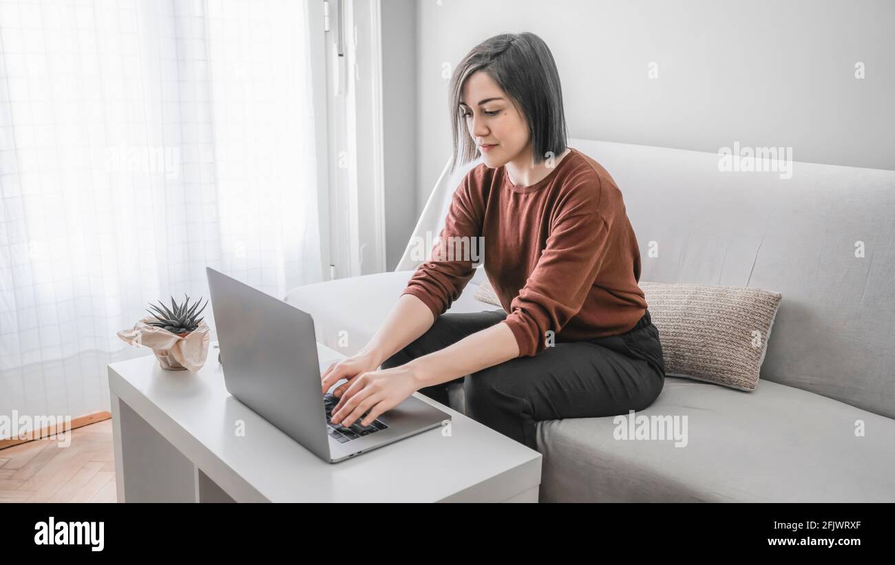 Woman smiling and working at home with her laptop sitting on a sofa ...