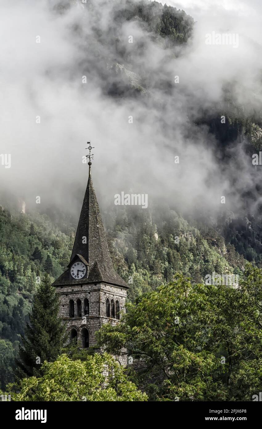bell tower of a church with a big clock in a mountain village in the Pyrenees with clouds behind it coming down the mountainside, church of Arties, Ll Stock Photo