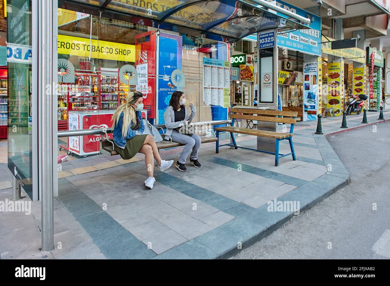 Just before curfew, young Turkish women wait for public transport while sitting on bus stop bench. Stock Photo