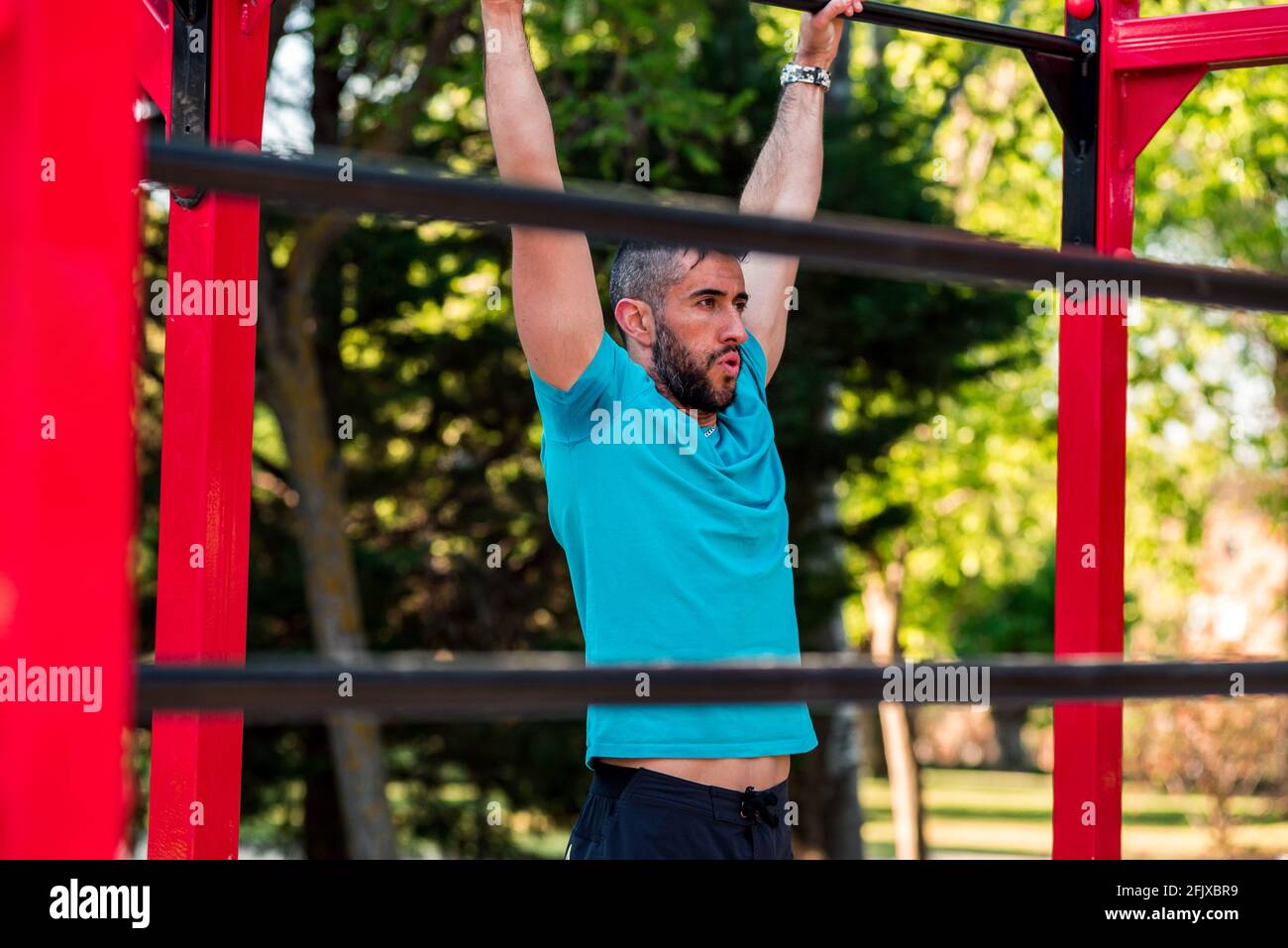 Dark-haired athlete with beard hanging from a calisthenics bar. View between bars. Outdoor crossfit concept. Stock Photo