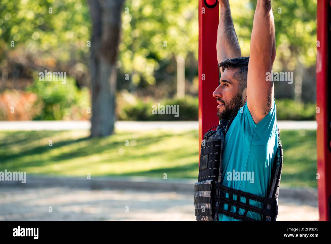 Bearded brunette man doing a barbell pull-up with weight vest. Outdoor fitness concept. Stock Photo