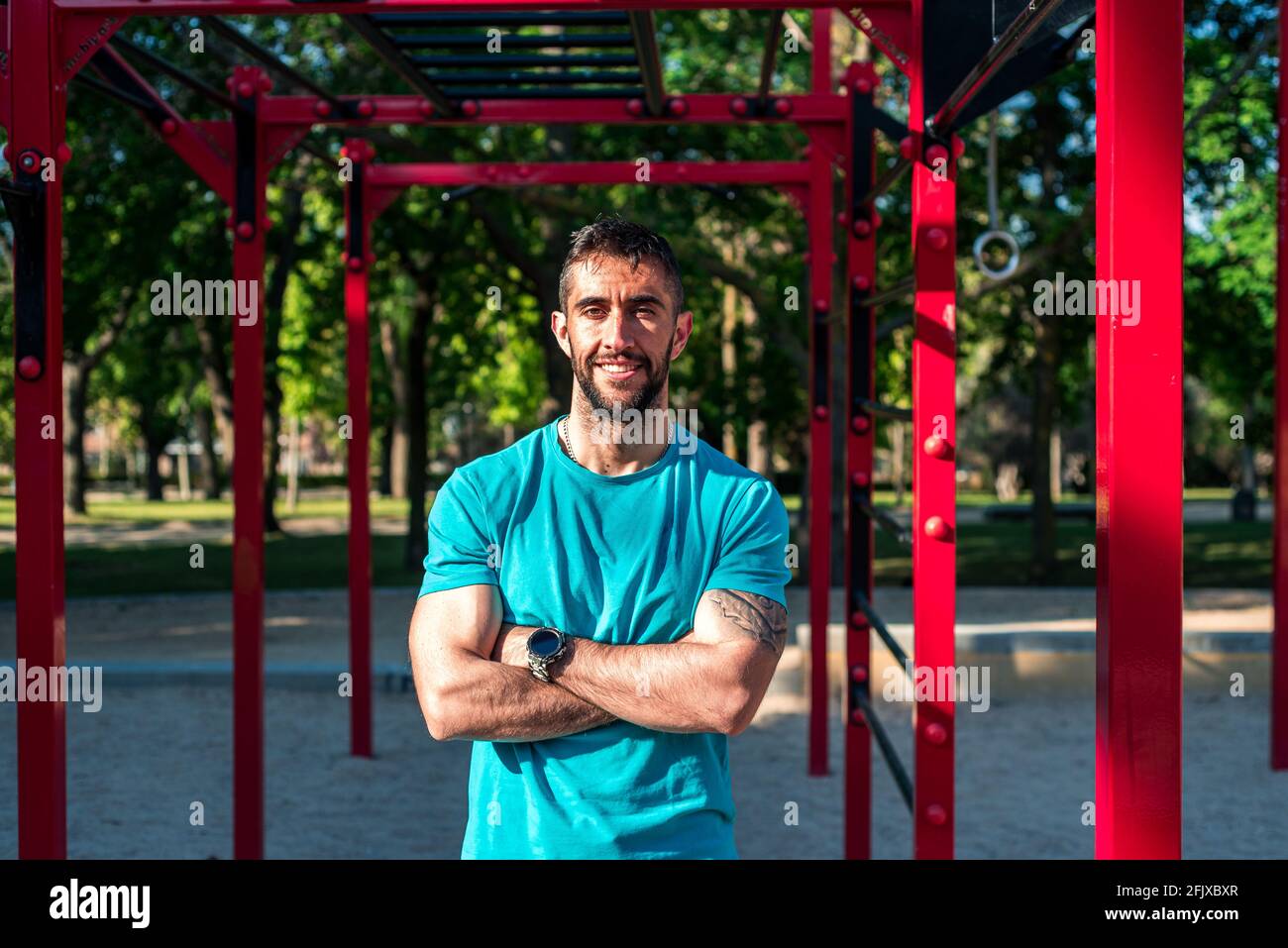 Portrait of a brunette bearded athlete with crossed arms. Red calisthenics bars in the background . Outdoor fitness concept. Stock Photo