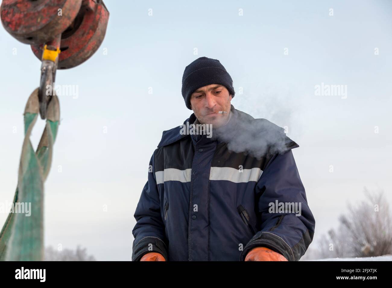 Portrait of a worker in a blue jacket and knitted hat at the hook of a truck crane with a ciga-rette in his mouth Stock Photo