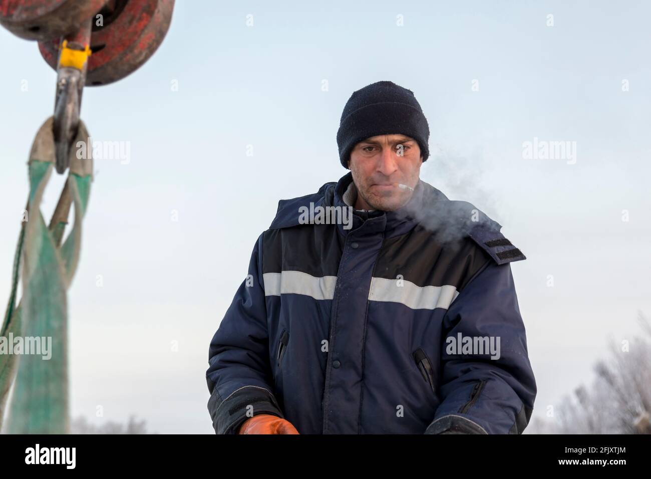Portrait of a worker in a blue jacket and knitted hat at the hook of a truck crane with a ciga-rette in his mouth Stock Photo