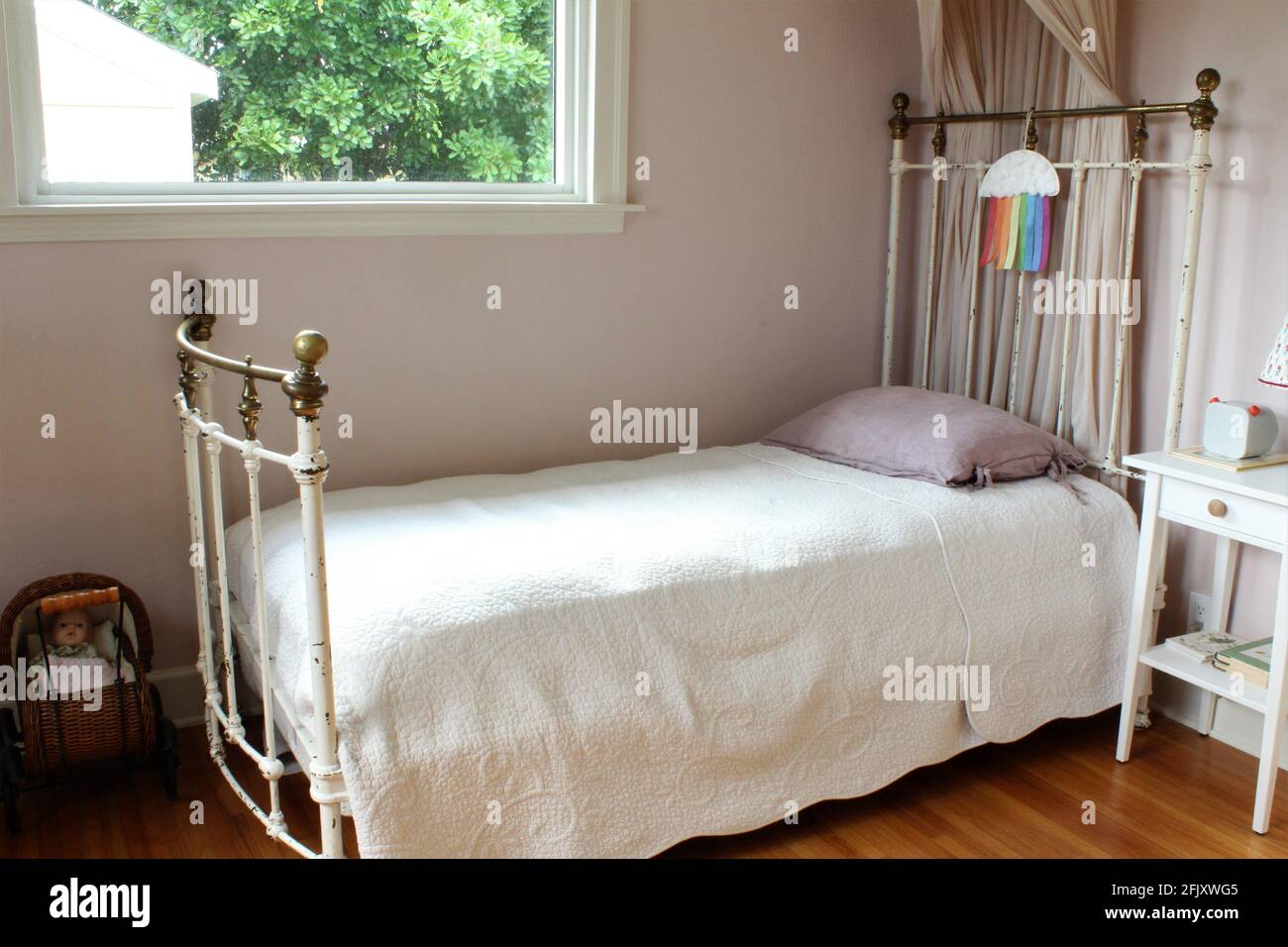 Girl's bedroom. Iron frame canopy bed with small desk lamp on a night stand. A small baby carriage is in the room for play. Big open space window Stock Photo