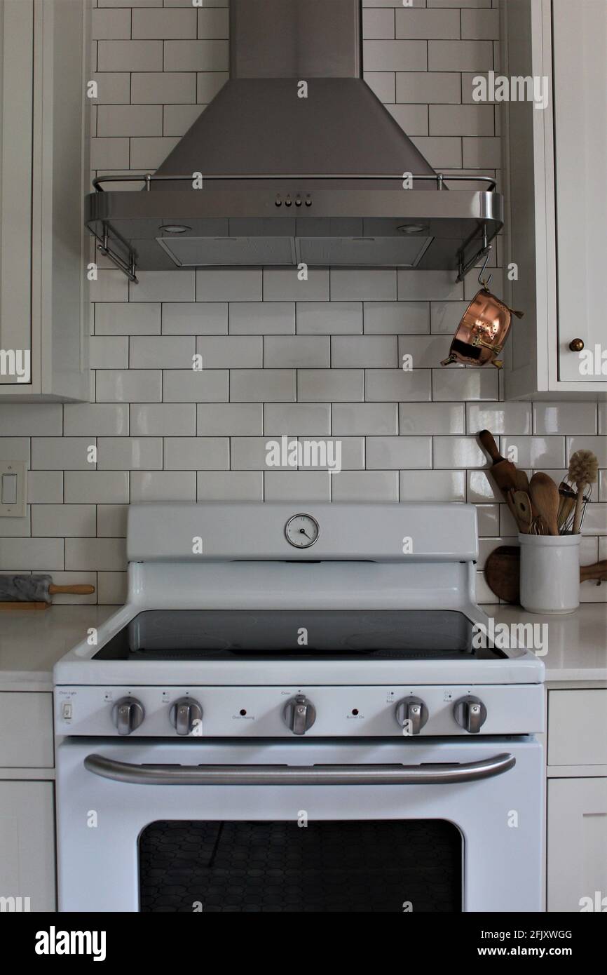 White Kitchen cupboard with white subway tile. Kitchen utensils in a bowl for cooking with a small vintage copper colander. Large kitchen hood exhaust Stock Photo