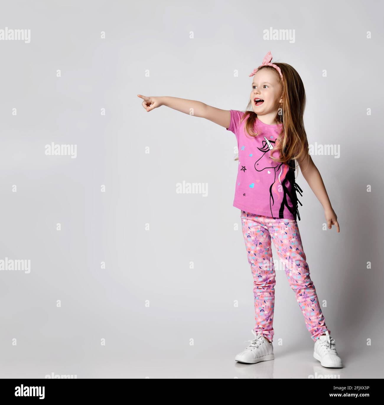 Happy screaming blonde kid girl in summer clothing colorful pants and t-shirt stands pointing with finger at copy space Stock Photo