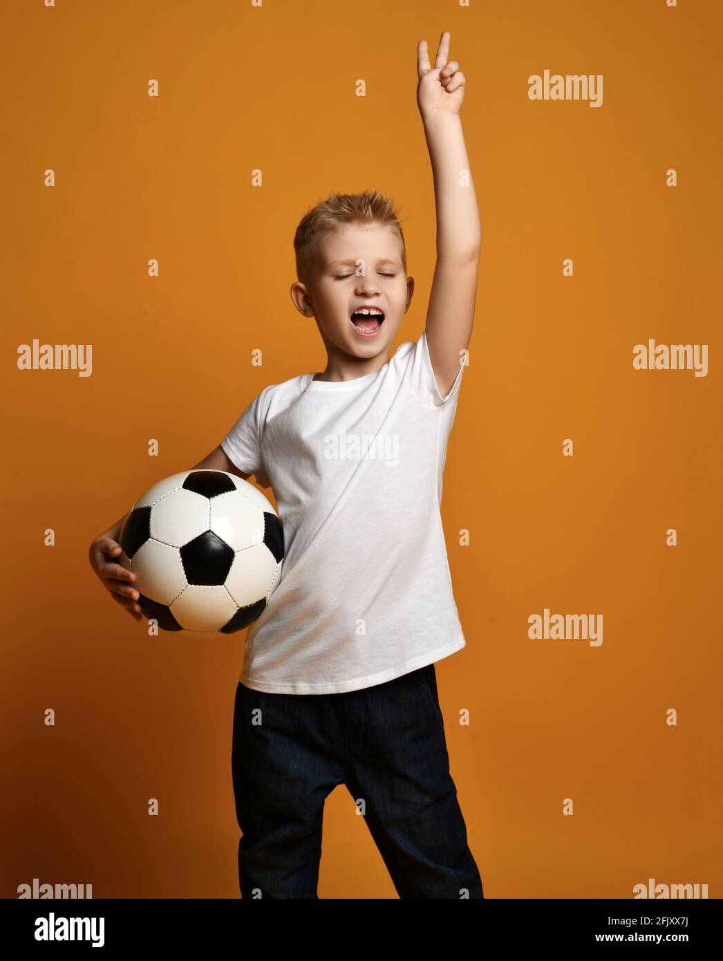 Happy kid boy in white blank t-shirt stands holding soccer ball in hand and gestures V victory sign, celebrates win Stock Photo