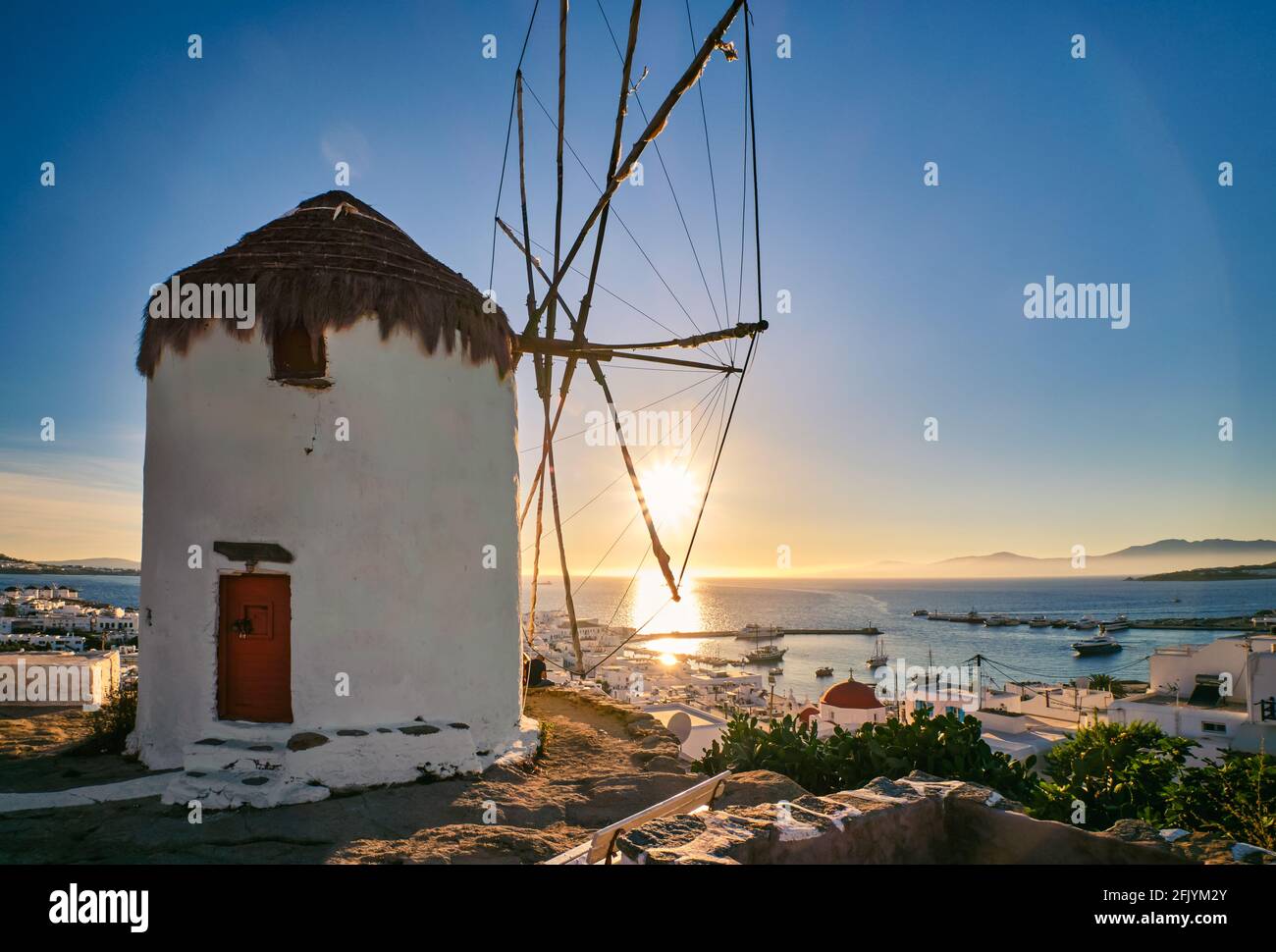 Romantic view of traditional Greek whitewashed windmill on hill. Chora town and harbor of Mykonos, Greece, low sun above sea horizon, sunset sky. Stock Photo