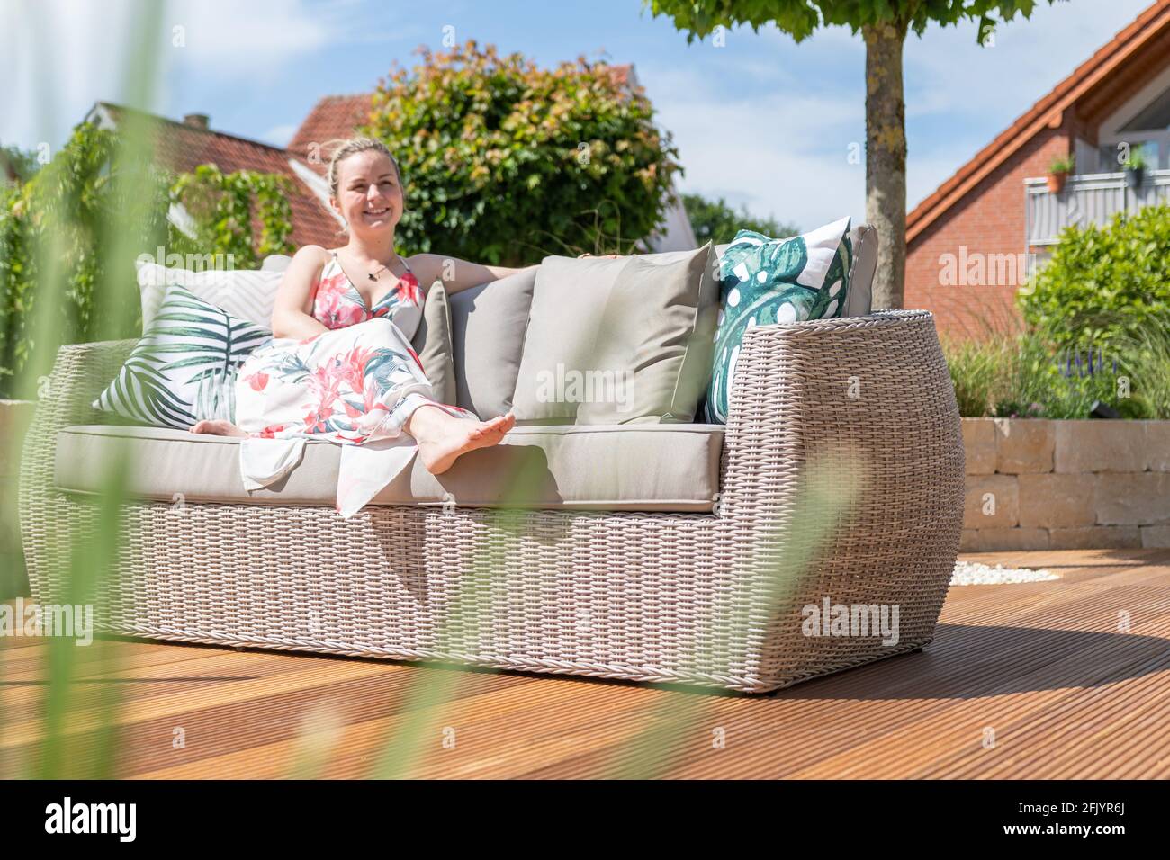 Attractive blonde woman and dog relaxing on a wooden terrace with green garden and modern outdoor furniture on a sunny day Stock Photo