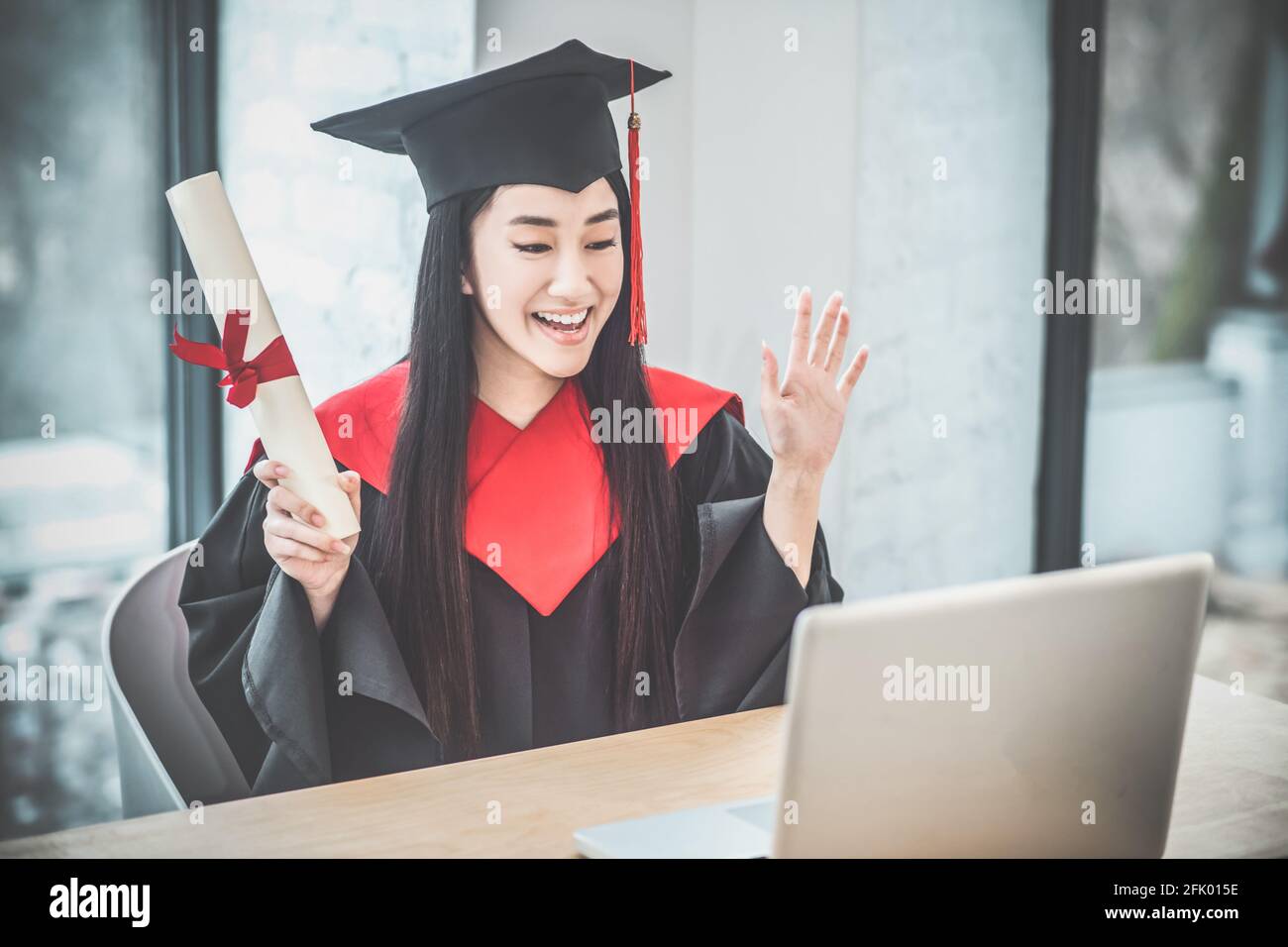 Cute asian smiling graduate holding her diploma and having a video call ...
