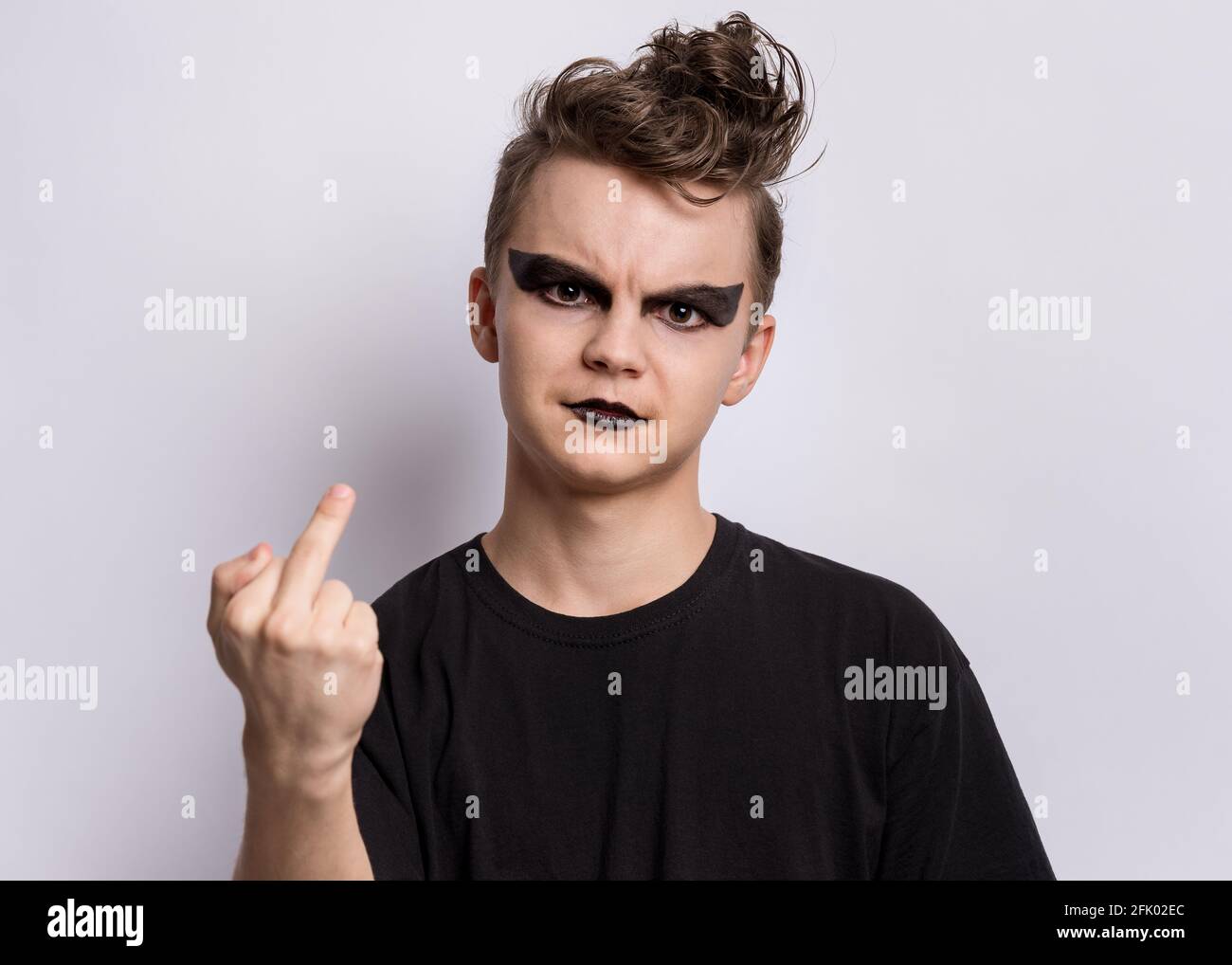 Crazy teen boy with spooking make-up making of punk goth dressed in black, showing middle finger on gray background. Problems of transitional age Stock Photo