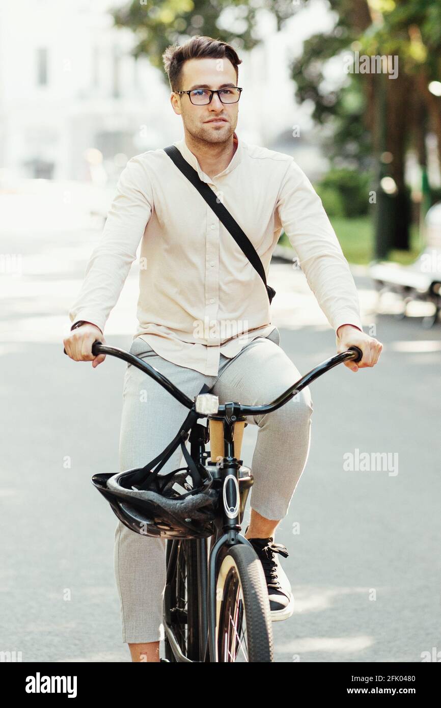 Modern office worker, personal eco transport in city and active lifestyle. Serious millennial attractive male in glasses with bag rides on bicycle in Stock Photo