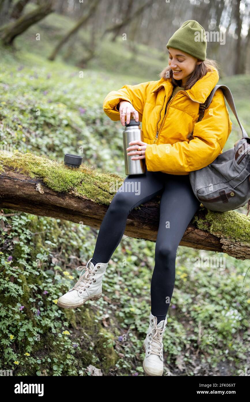 Young woman in hiking colorful clothes and backpack sitting on a tree and  drinking hot tea from a thermos. Enjoys the freshness of the air and the  tranquility of nature in the