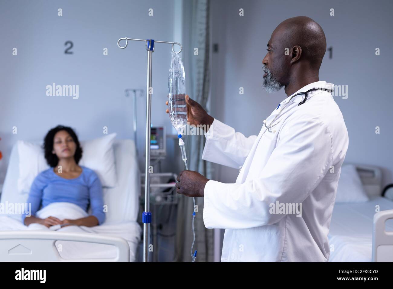 African american male doctor preparing iv drip bag for mixed race woman sitting up in hospital bed Stock Photo