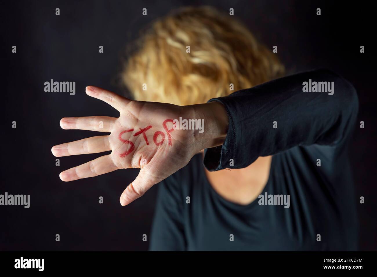 A blonde woman dressed in black covers her face with a hand on which she has written the word stop with a red marker, to stop violence against women Stock Photo