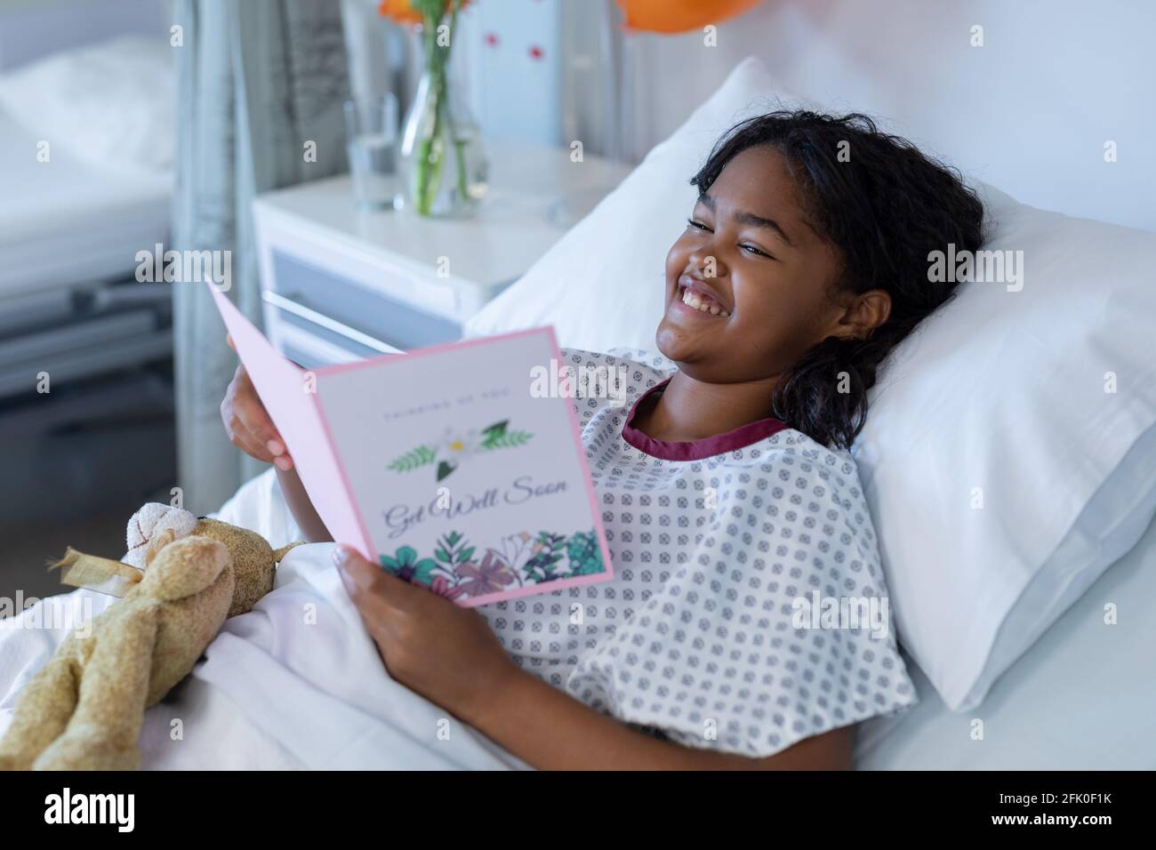 Smiling sick mixed race girl lying in hospital bed reading get well greetings card and holding teddy Stock Photo