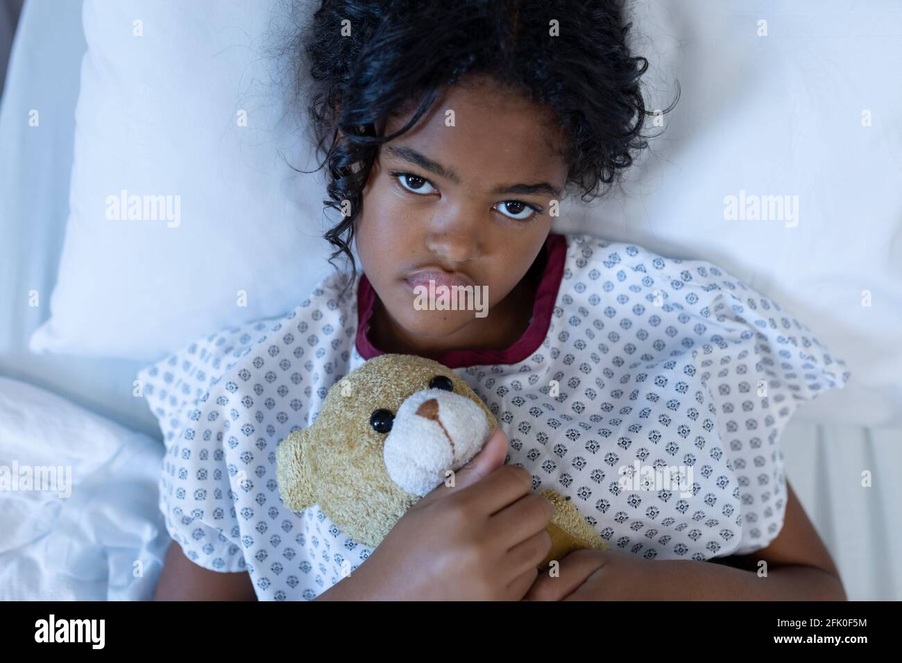 Portrait of sick mixed race girl lying in hospital bed holding teddy bear Stock Photo