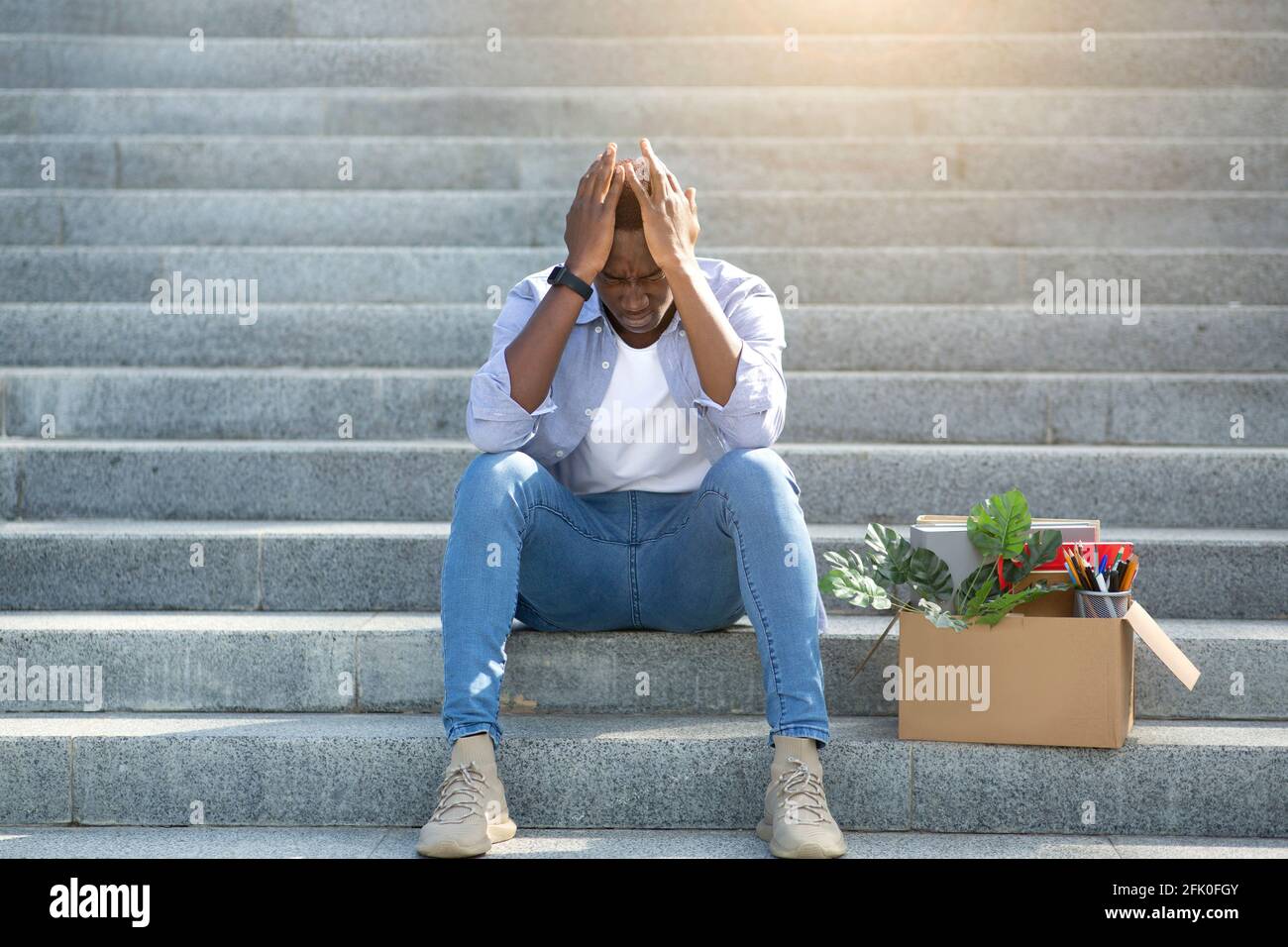 Economic crisis and bankruptcy. Desperate black man with personal belongings sitting on stairs after losing work, outside Stock Photo