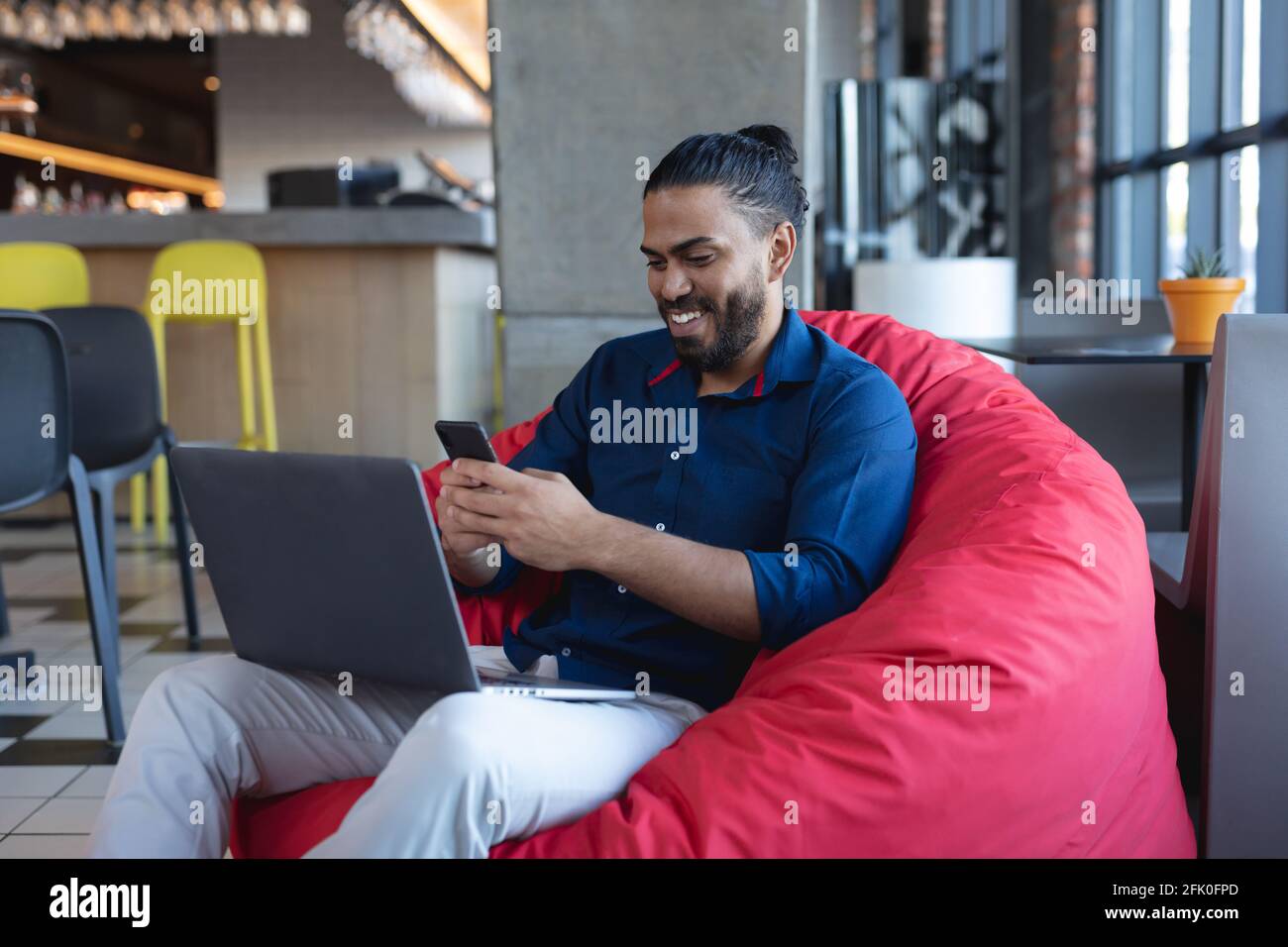 Smiling mixed race man sitting, using smartphone and laptop in cafe Stock Photo