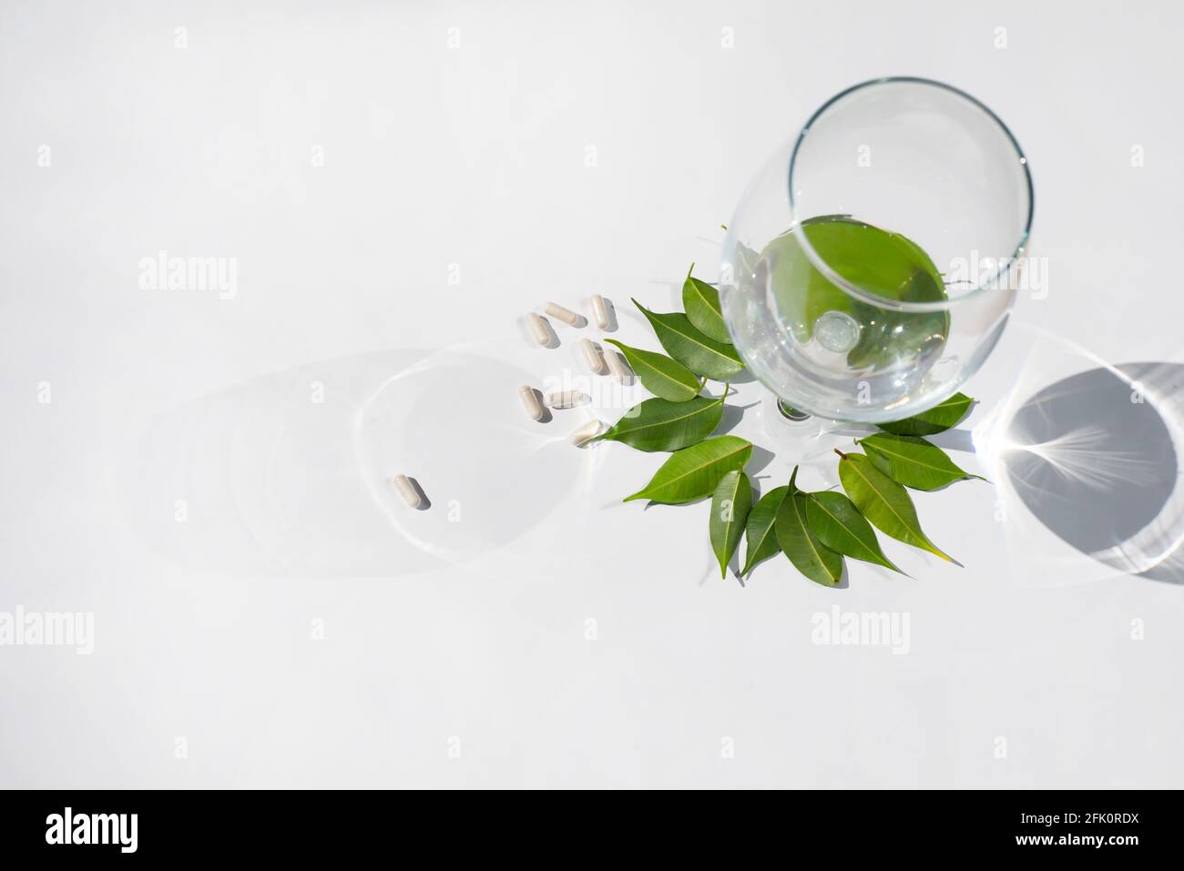 Glass wine goblet filled with water. Pill capsules on a white background and green leaves. The topic of nutraceuticals and supplementation.  Stock Photo