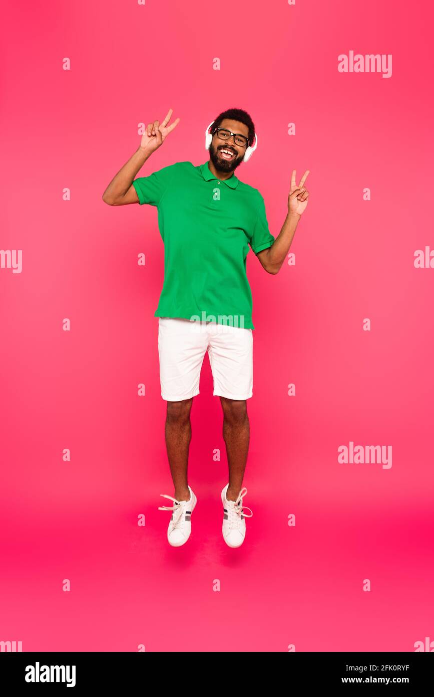 full length of happy african american man in headphones showing peace sign and jumping on pink Stock Photo