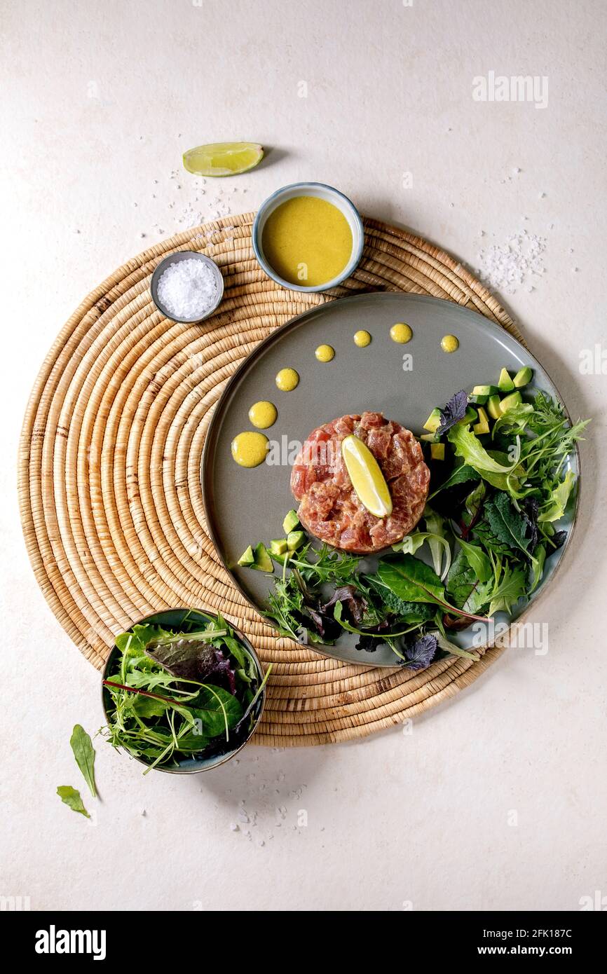 Tuna tartare with green salad, lime, avocado and mustard sauce serving on ceramic plate on straw napkin over white texture background. Flat lay, copy Stock Photo