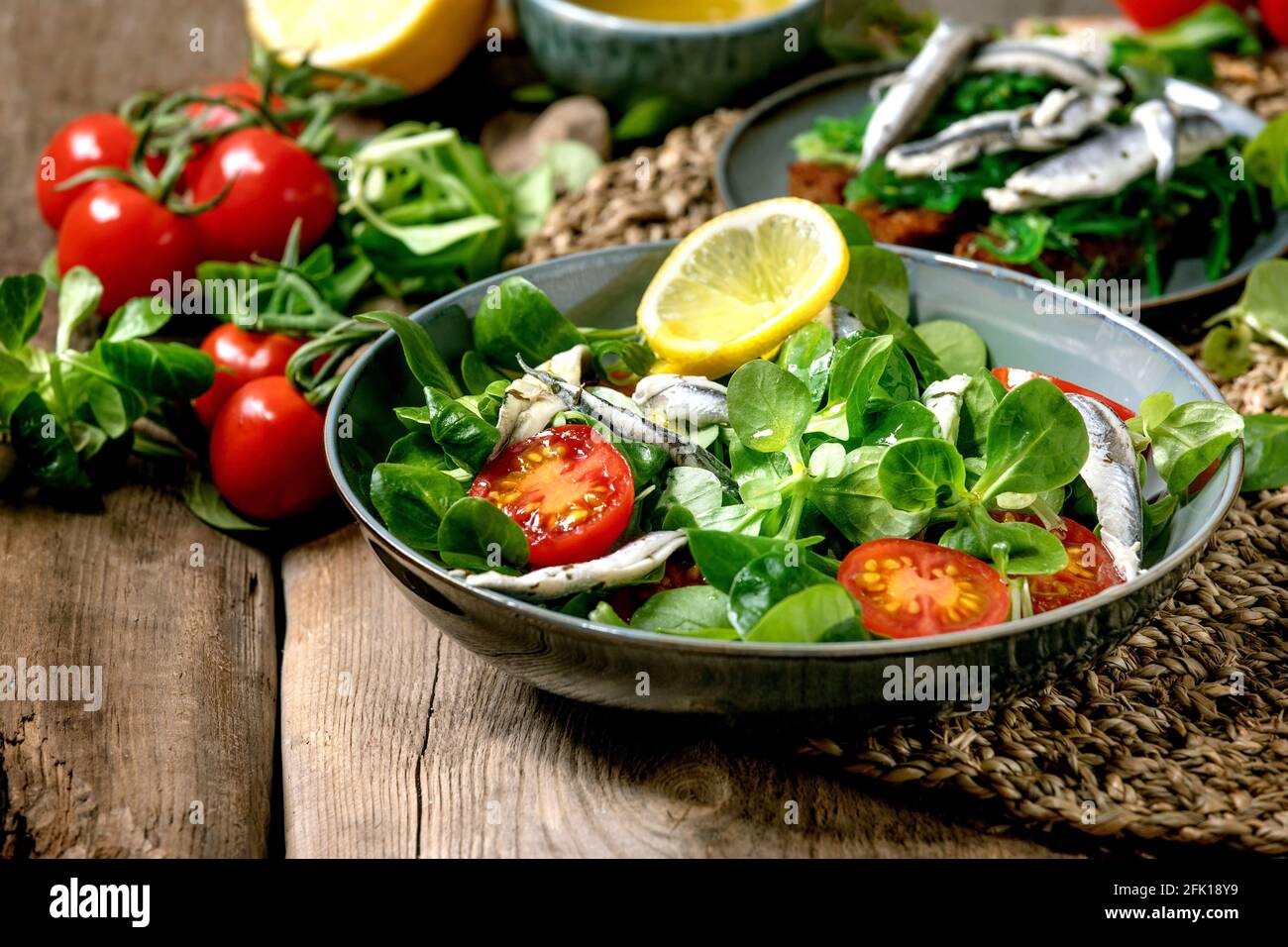 Green field salad with pickled anchovies or sardines fillet, and cherry tomatoes, served in blue bowl with lemon and olive oil on straw napkin over ol Stock Photo