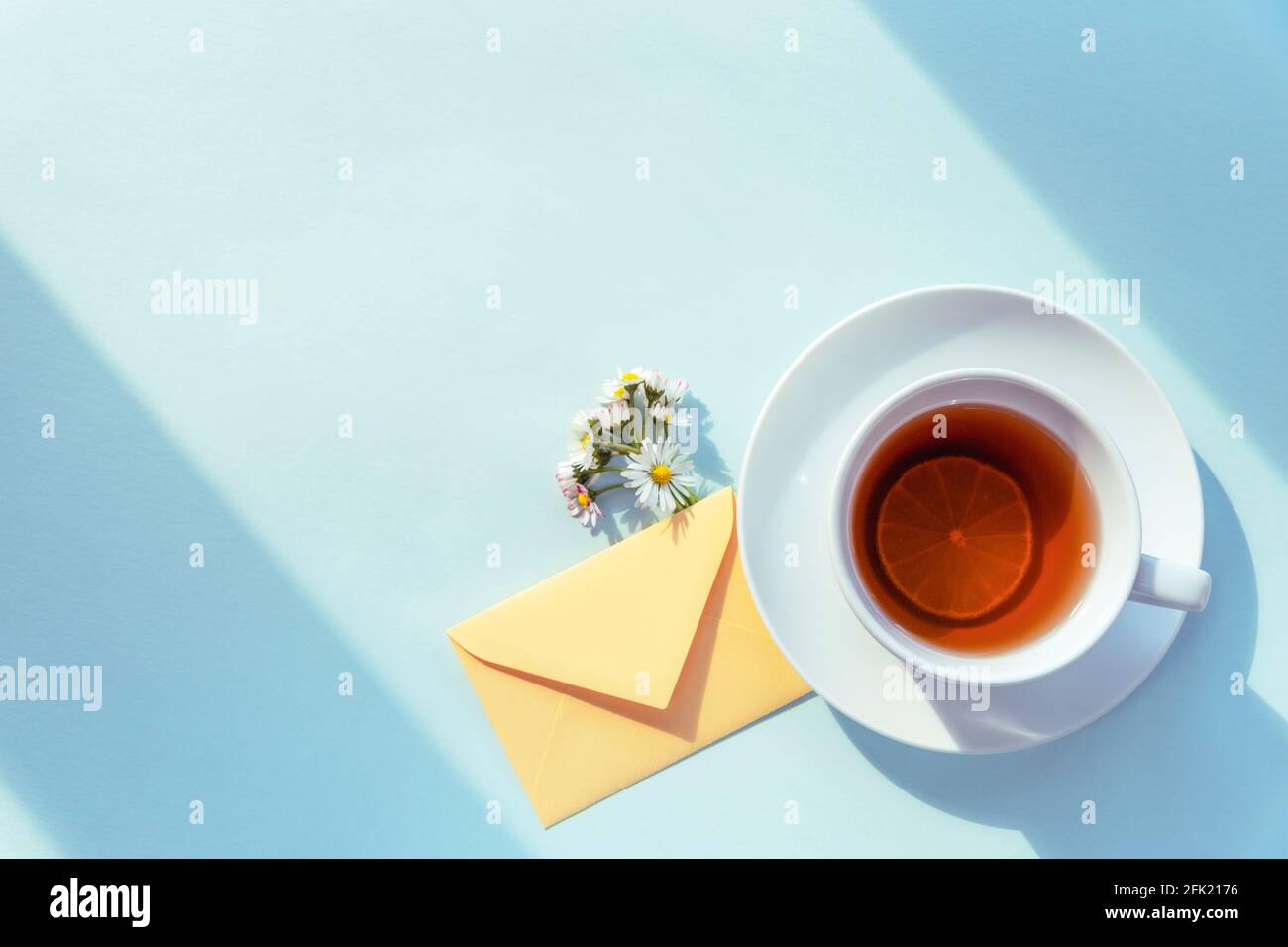 A cup of tea, an envelope and a bouquet of daisies in sunlight on blue background. Top view, flat lay. Stock Photo