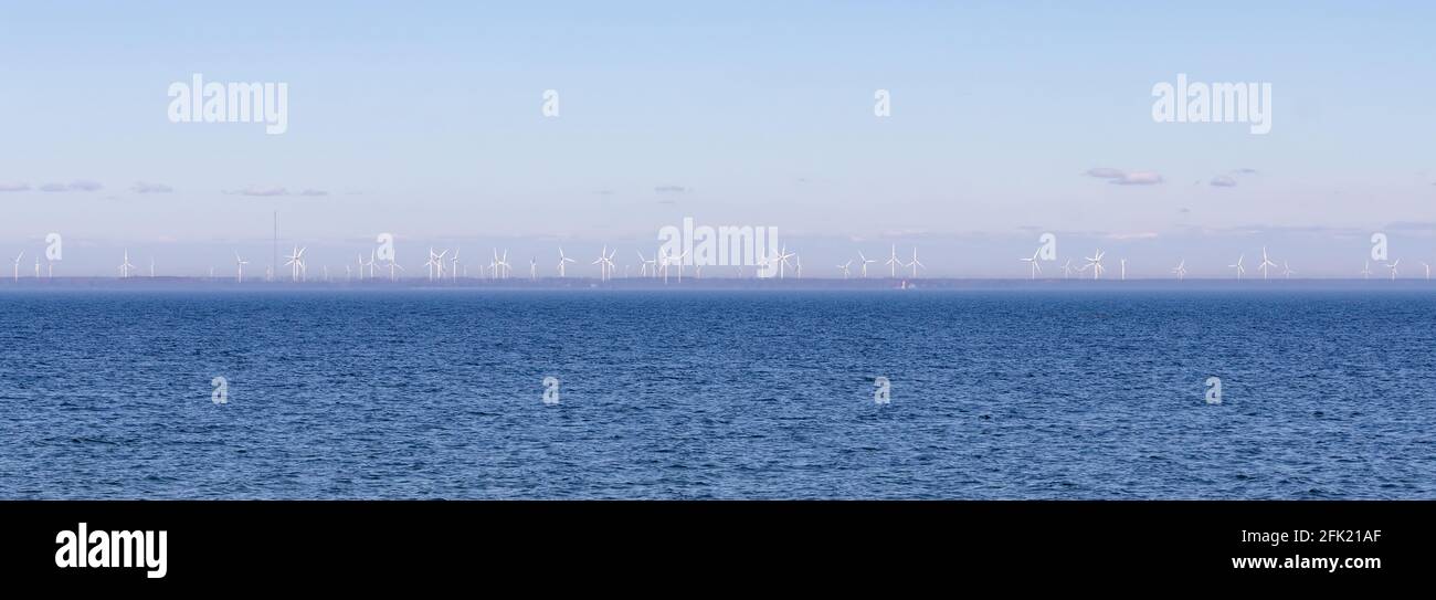 Panoramic view of open water and a wind farm on shore full of wind turbines. Light hazy blue sky over dark blue water. Lake Ontario, Canada Stock Photo