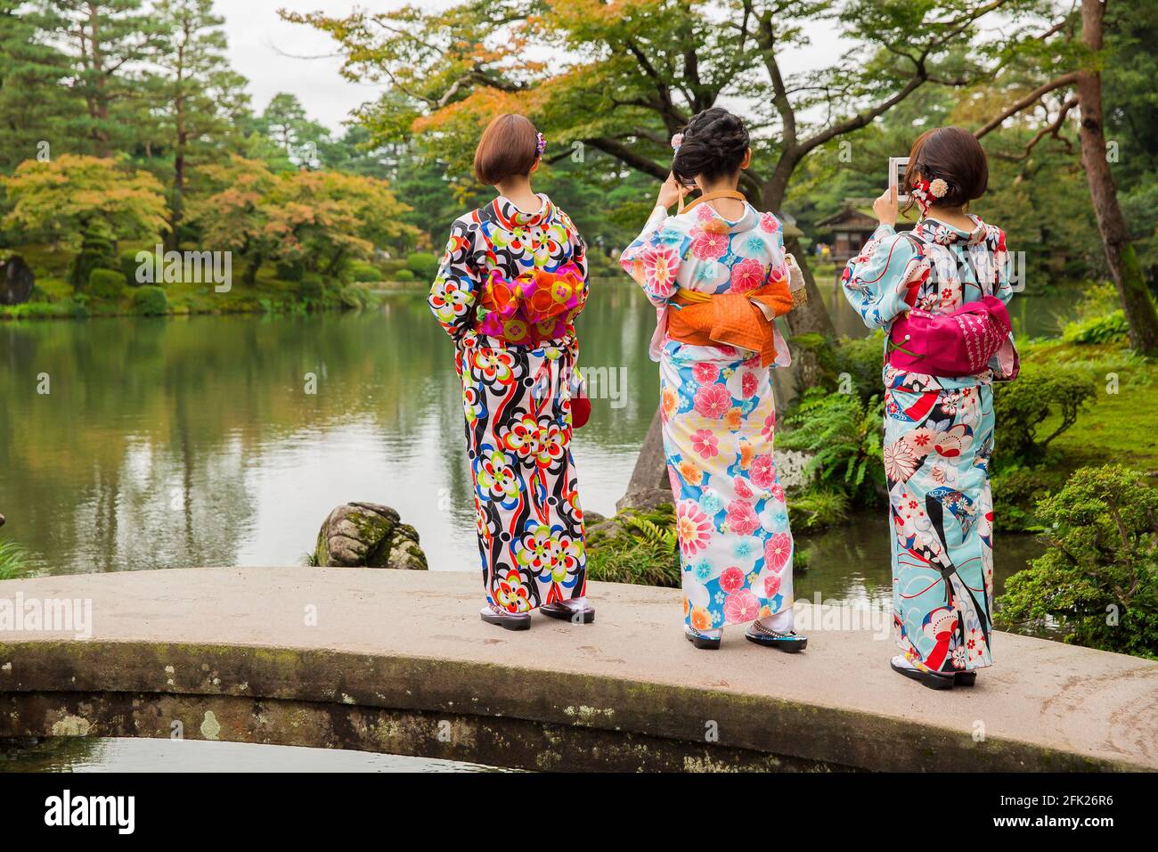 Young Japanese girls, ladies, women wearing bright colourful kimono dress. Geisha maiko standing on bridge over lake. Kenrokuen Garden, Kanazawa Japan Stock Photo