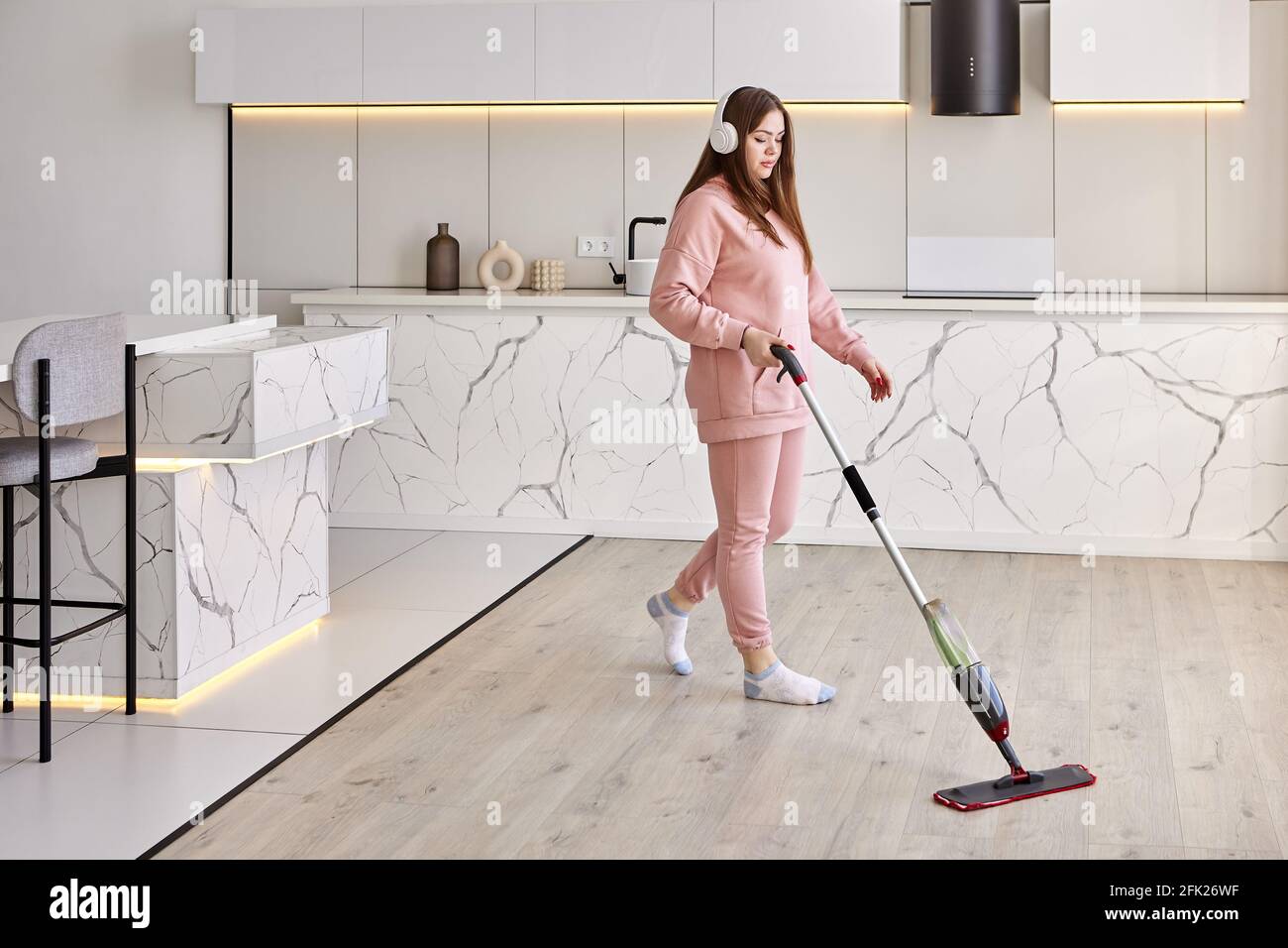 Washing floor using flat mop with microfiber pad and spray bottle, young white woman tidies up kitchen. Stock Photo