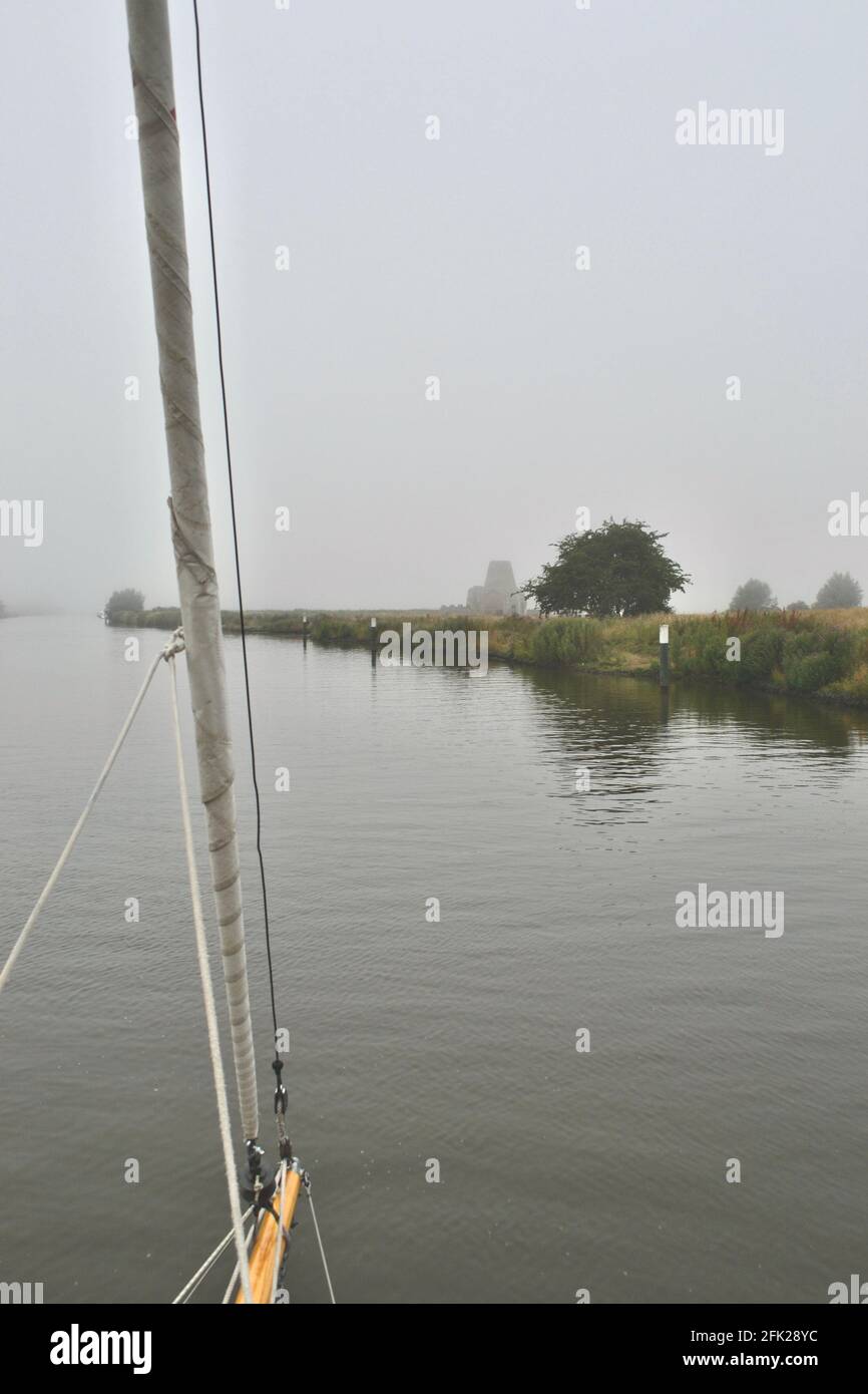 Sailing into the mist: the wooden bowsprit of boat (sailing yacht) approaching a misty, reedlined shore with a tree and a building in the fog in the d Stock Photo