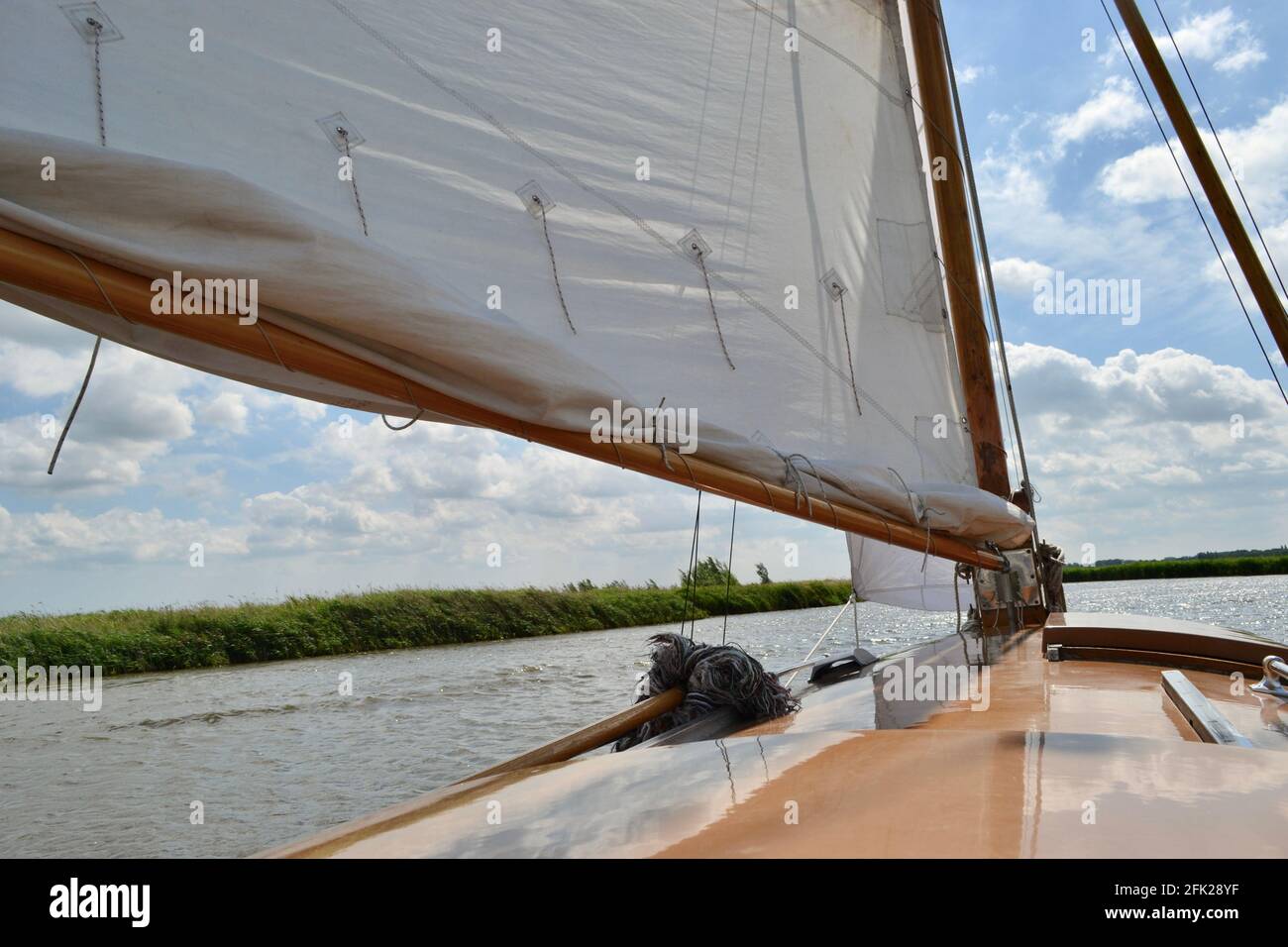 Looking forward along the cabin top of a traditional sailing yacht: a brown boat with white sails, on a summer's day, sailing along a river (Norfolk B Stock Photo