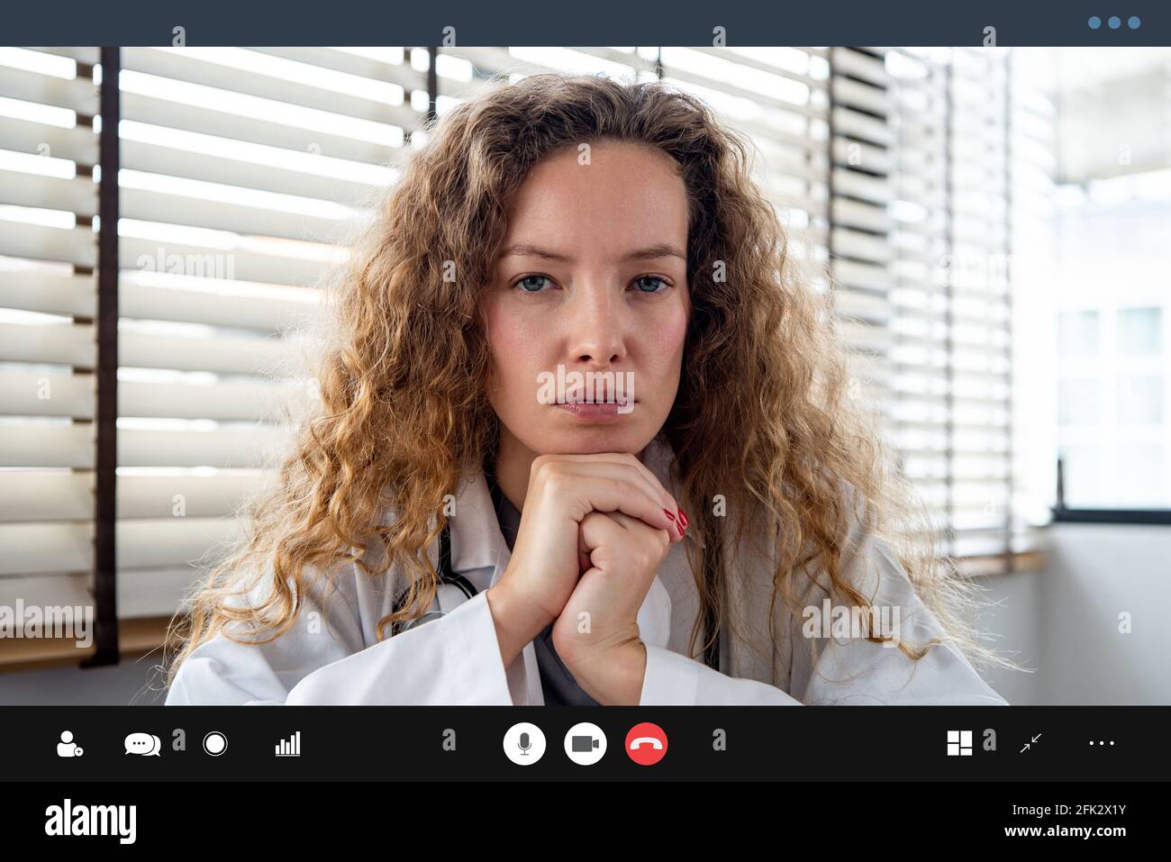 Serious female doctor carefully listening and keeping hands under chin while making medical conference video call from hospital Stock Photo