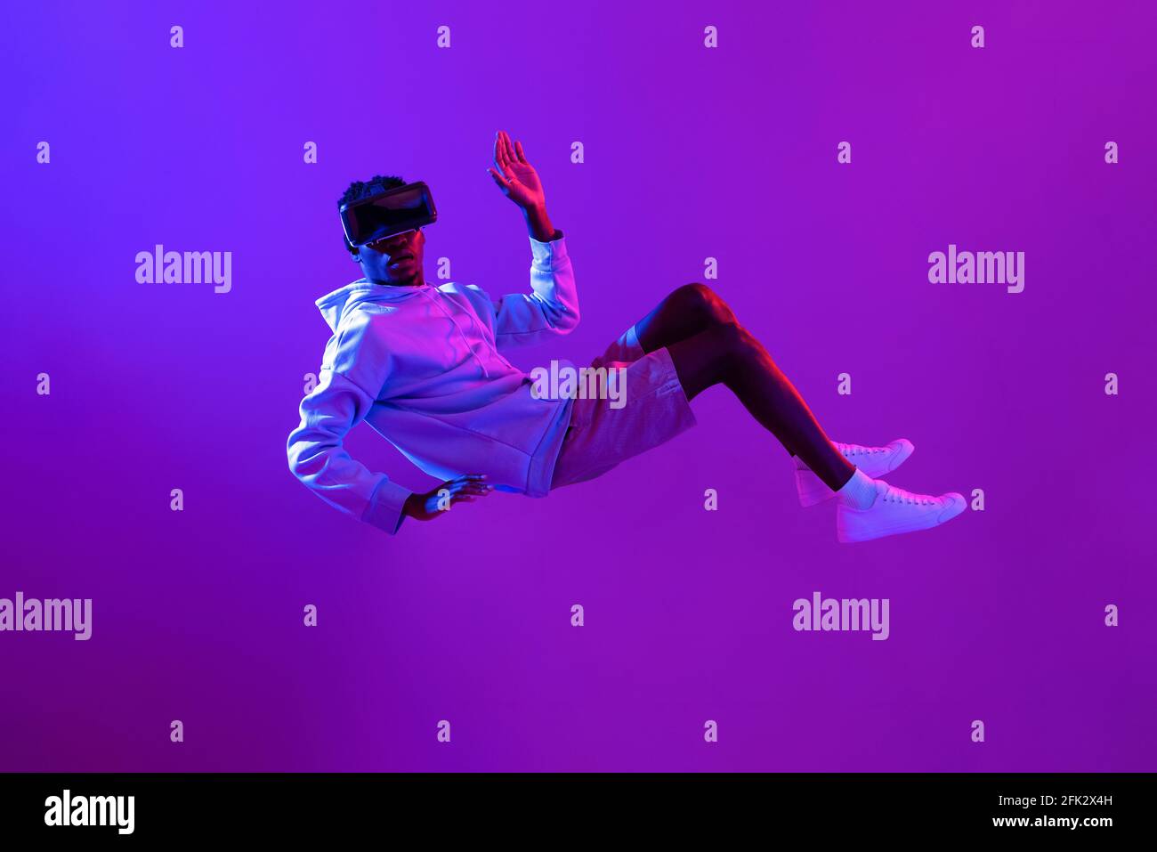 Amazed young African man wearing VR glass headset levitating in the air on futuristic purple cyberpunk neon light background Stock Photo
