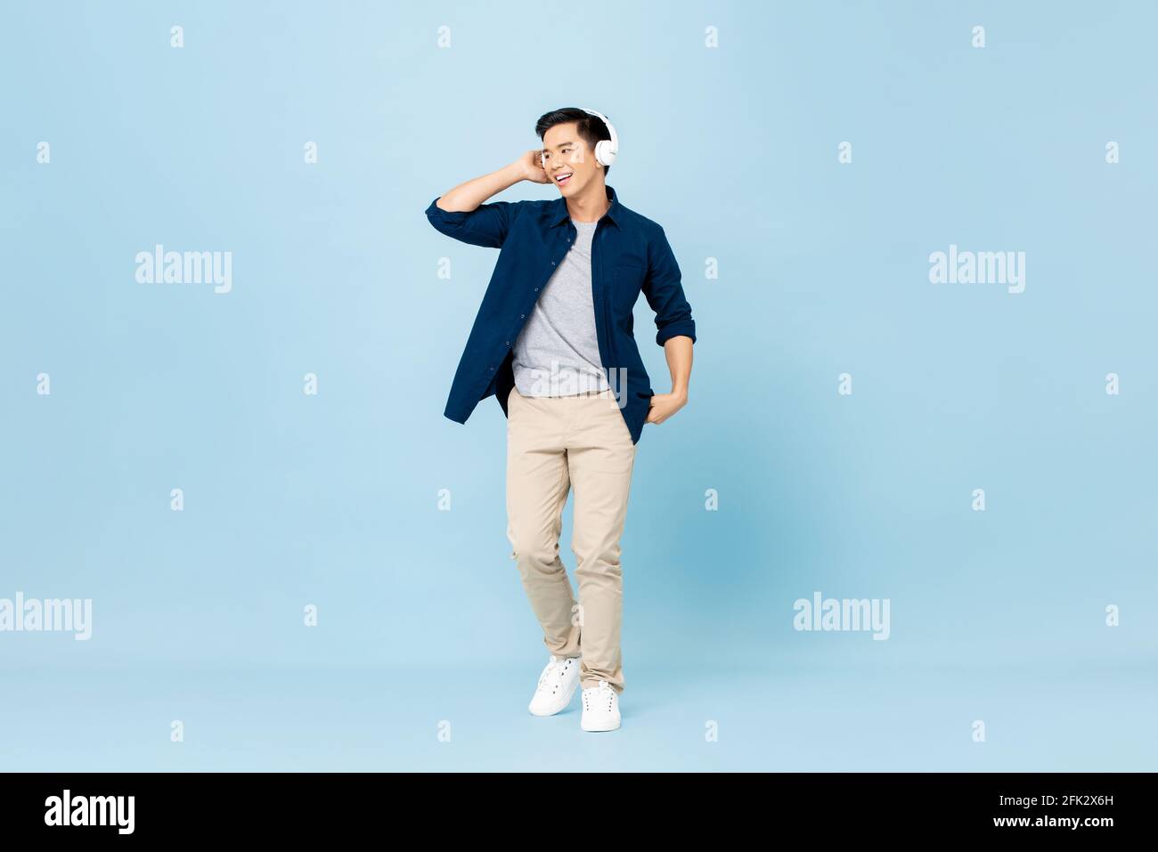 Full body portrait of smiling young handsome Asian man listening to music with wireless headphones isolated on light blue studio background Stock Photo