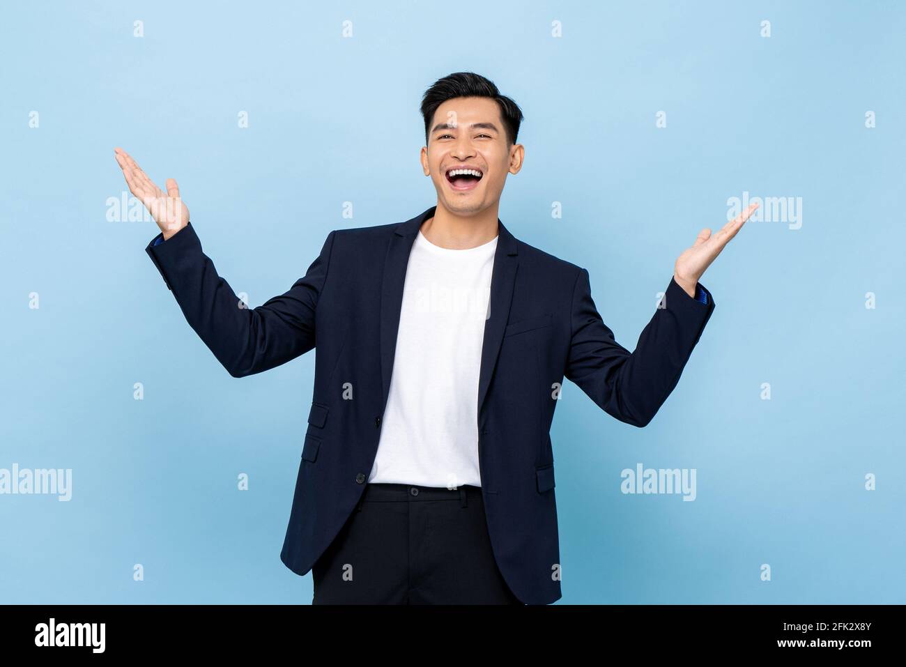 Surprised worry free handsome Asian man in semi formal clothes smiling and opening hands in light blue isolated studio background Stock Photo