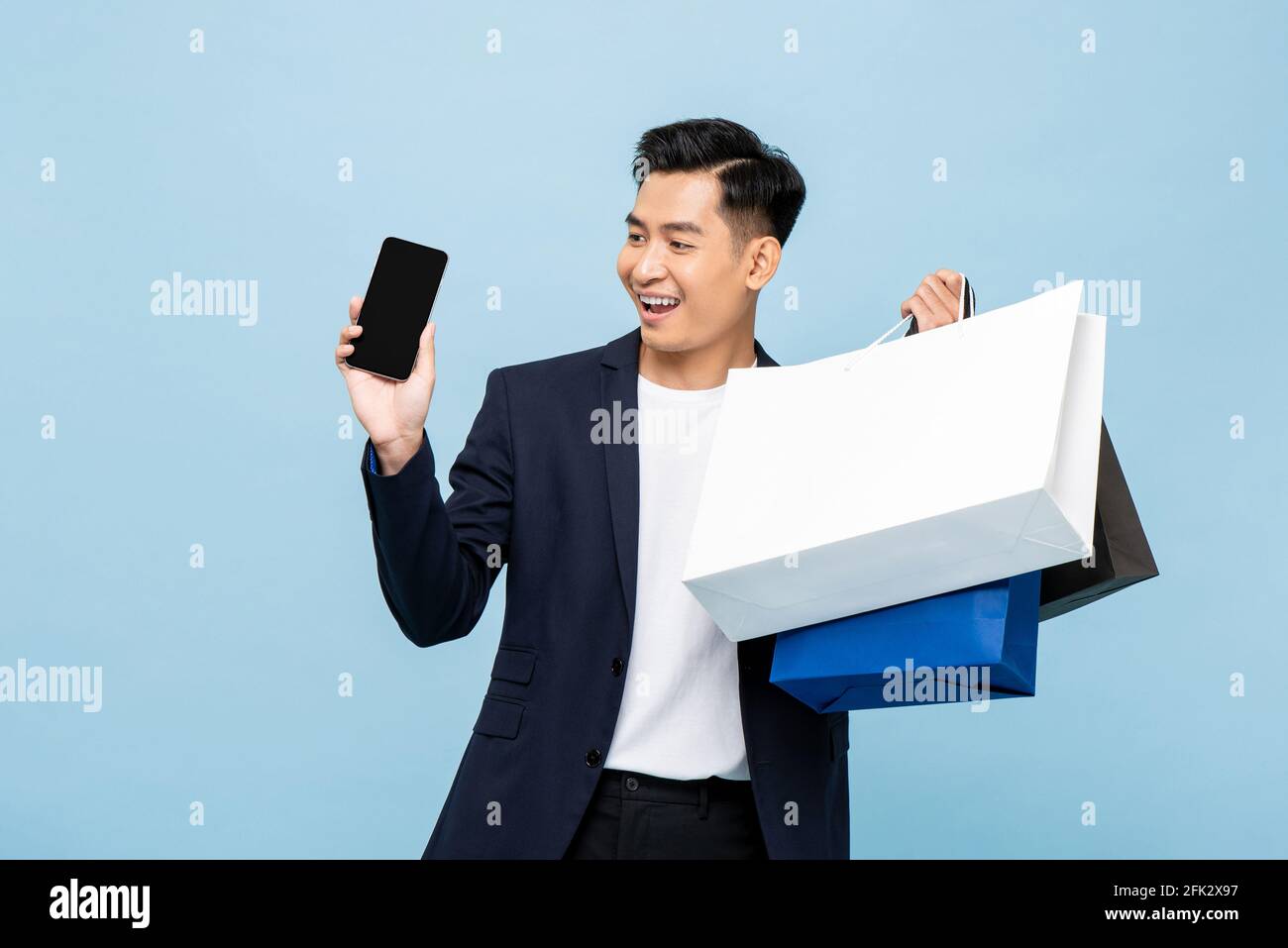 Young handsome southeast Asian man holding mobile phone and shopping bags in light blue studio isolated background Stock Photo