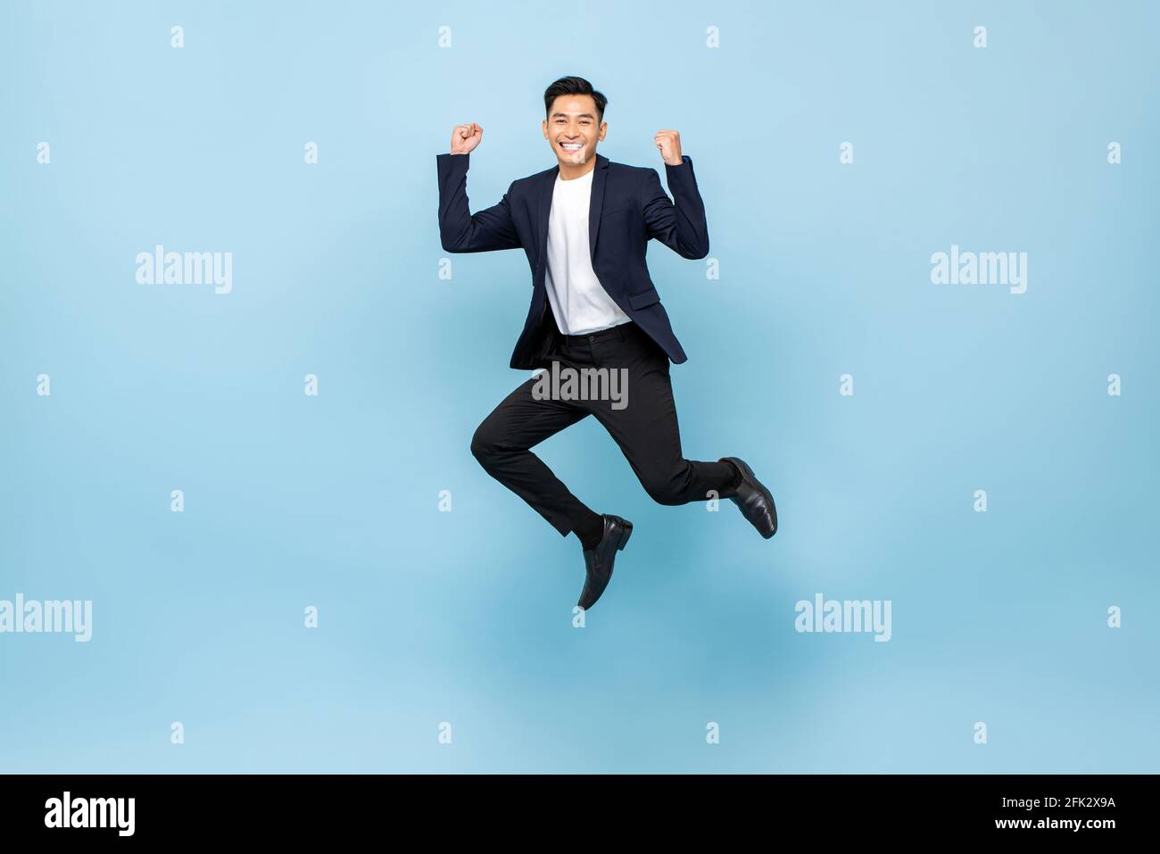 Full lenght portrait of smiling handsome Asian man jumping and raising  his fists on isolated light blue studio background Stock Photo