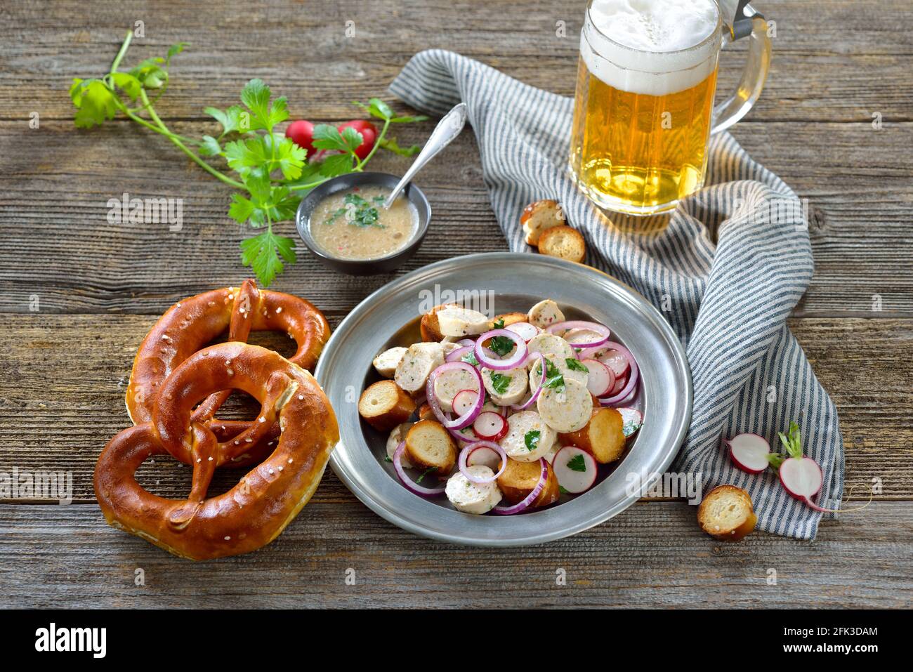 Hearty Bavarian salad with cut veal sausages, roasted pretzel slices on a tin plate, served with a mustard dressing and a mug of lager beer Stock Photo