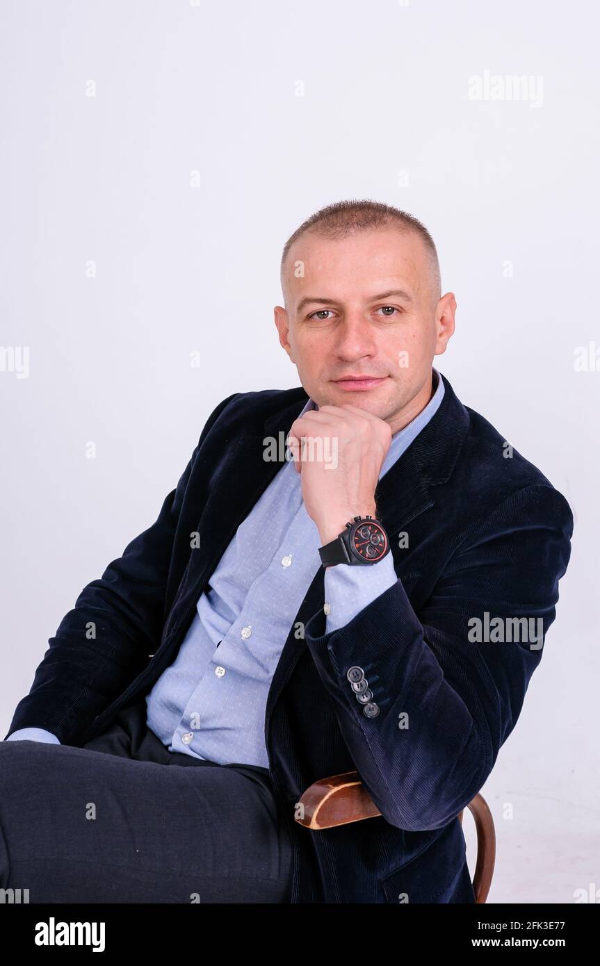 A man in a blue shirt, black trousers and a blue jacket sits on a stool on a white background Stock Photo