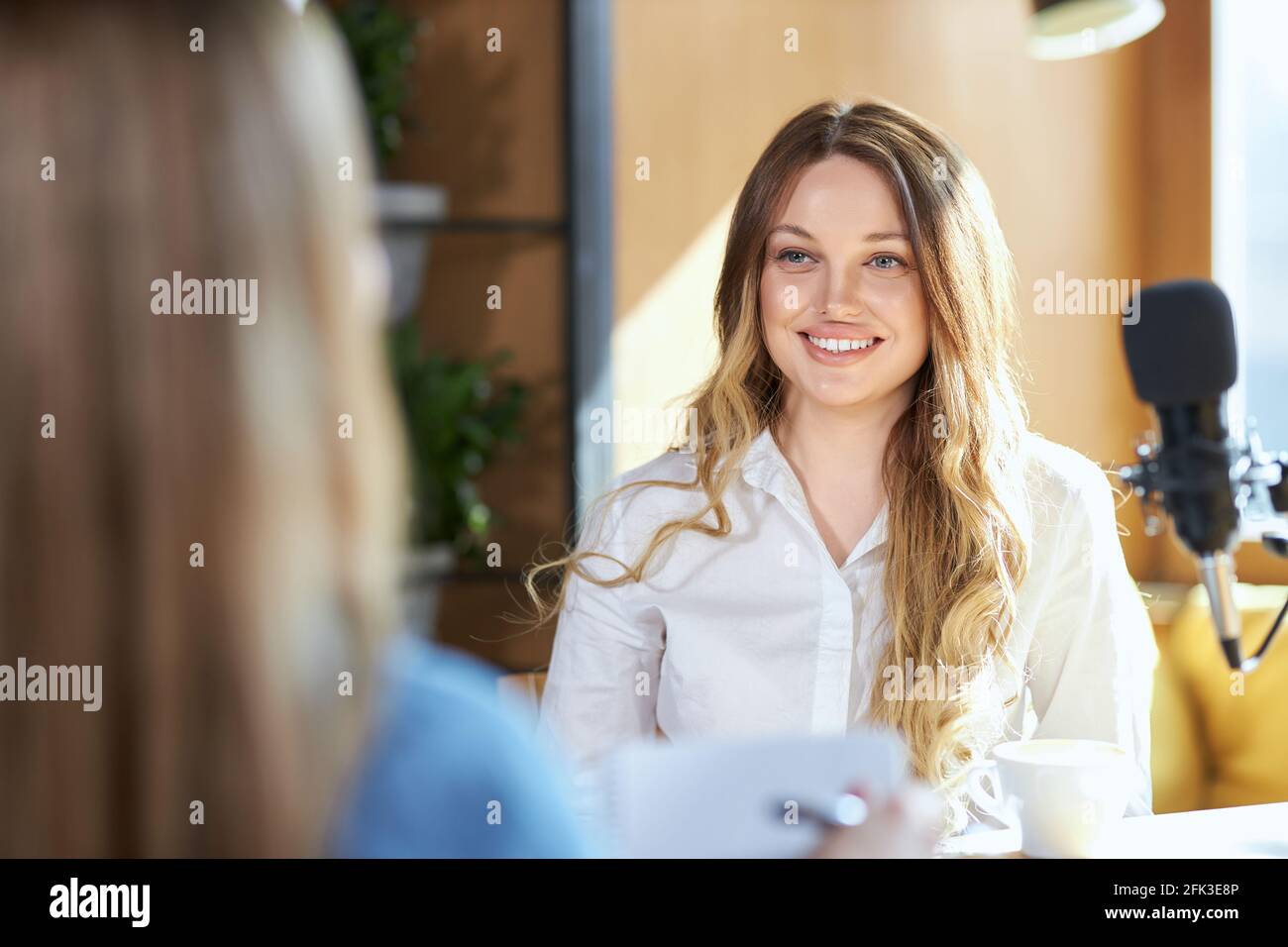 Front view portrait of smiling beautiful young woman giving interview in modern microphone with tasty coffee in cafe. Concept of process communicating about different topics with good mood.  Stock Photo