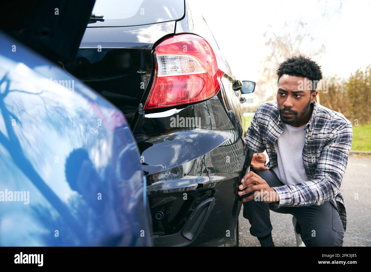 Angry young man standing by vandalised car in car park reporting damage to insurance company using mobile phone Stock Photo