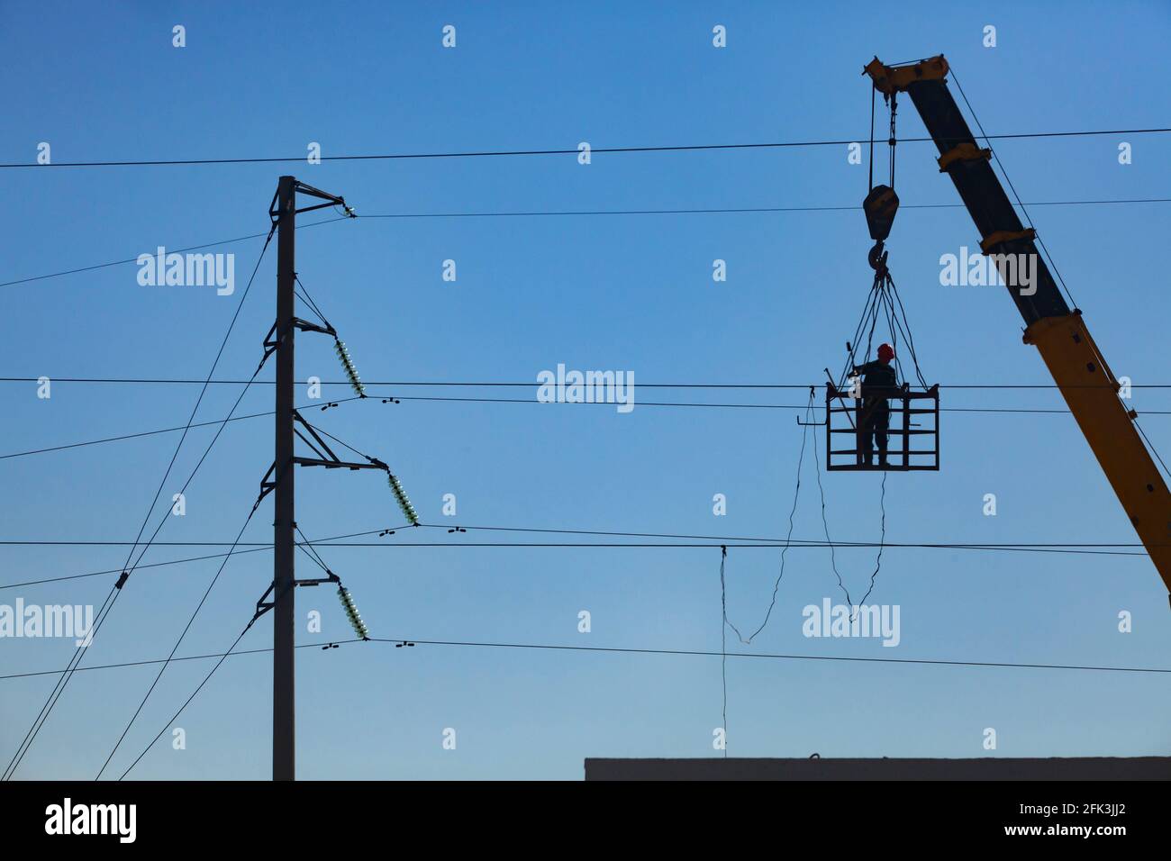 Silhouette of worker on the crane cradle. Repairing electric power line. Blue sky background. Stock Photo
