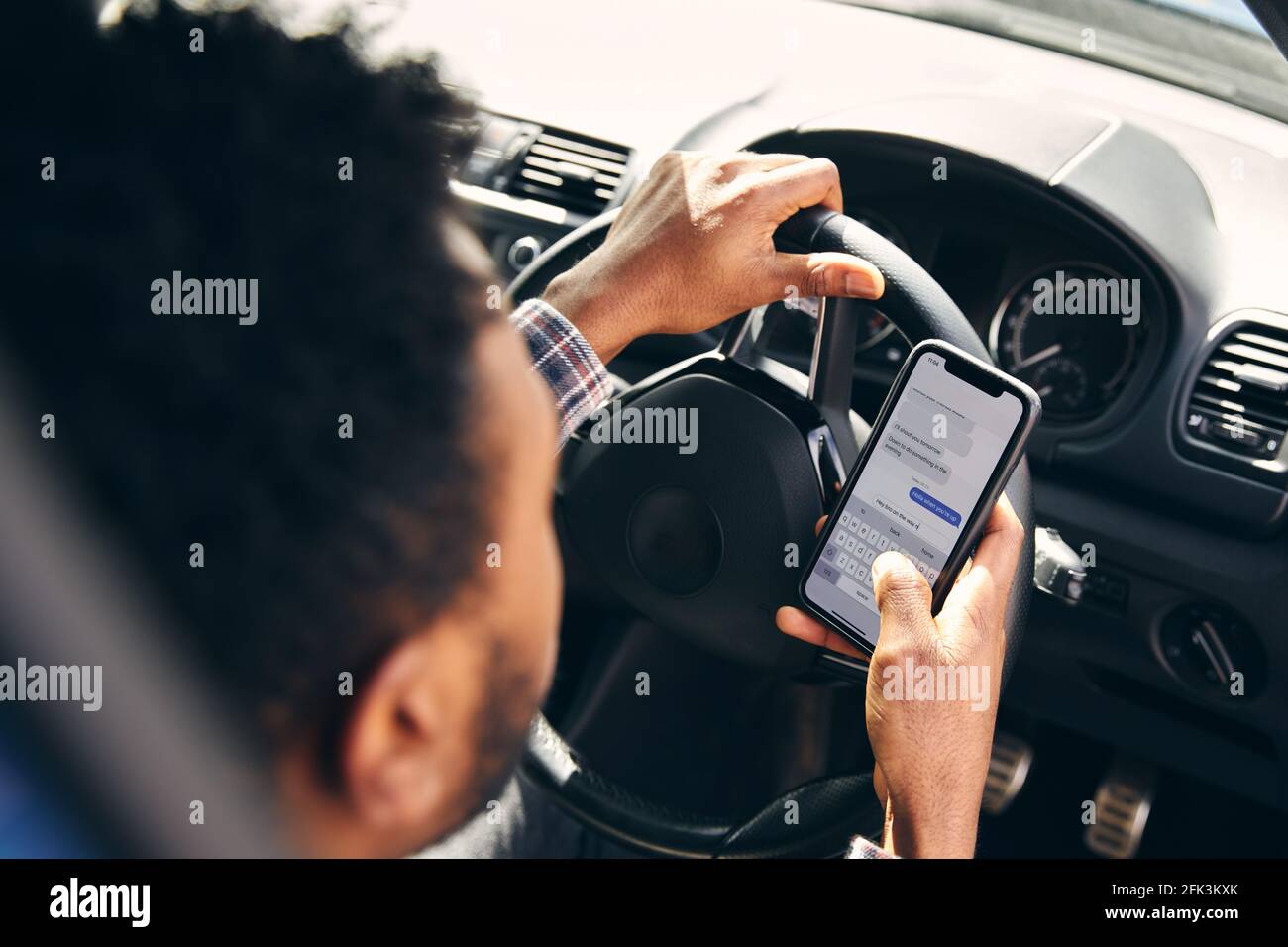 Over the shoulder view of young man driving dangerously texting on mobile phone with one hand on steering wheel Stock Photo