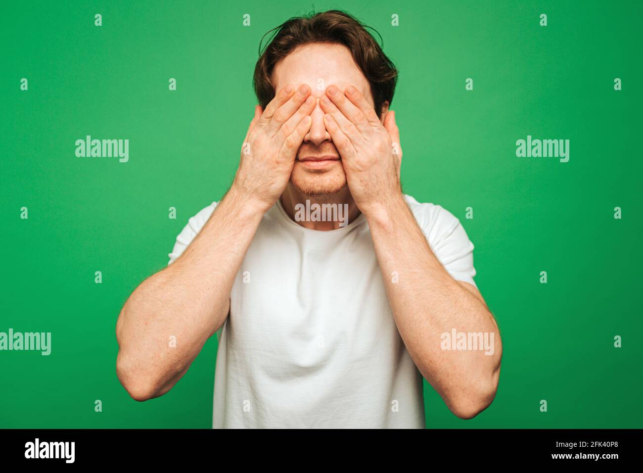 Young man covers eyes up with hands, isolated on green background Stock Photo