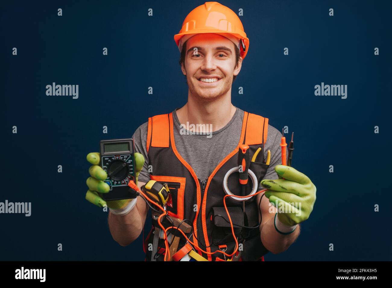 Happy builder is holding digital multimetert in gloves, isolated over blue background Stock Photo