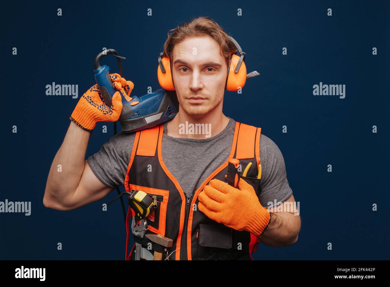 Young builder is serious while having equipment in arm, isolated over blue background Stock Photo