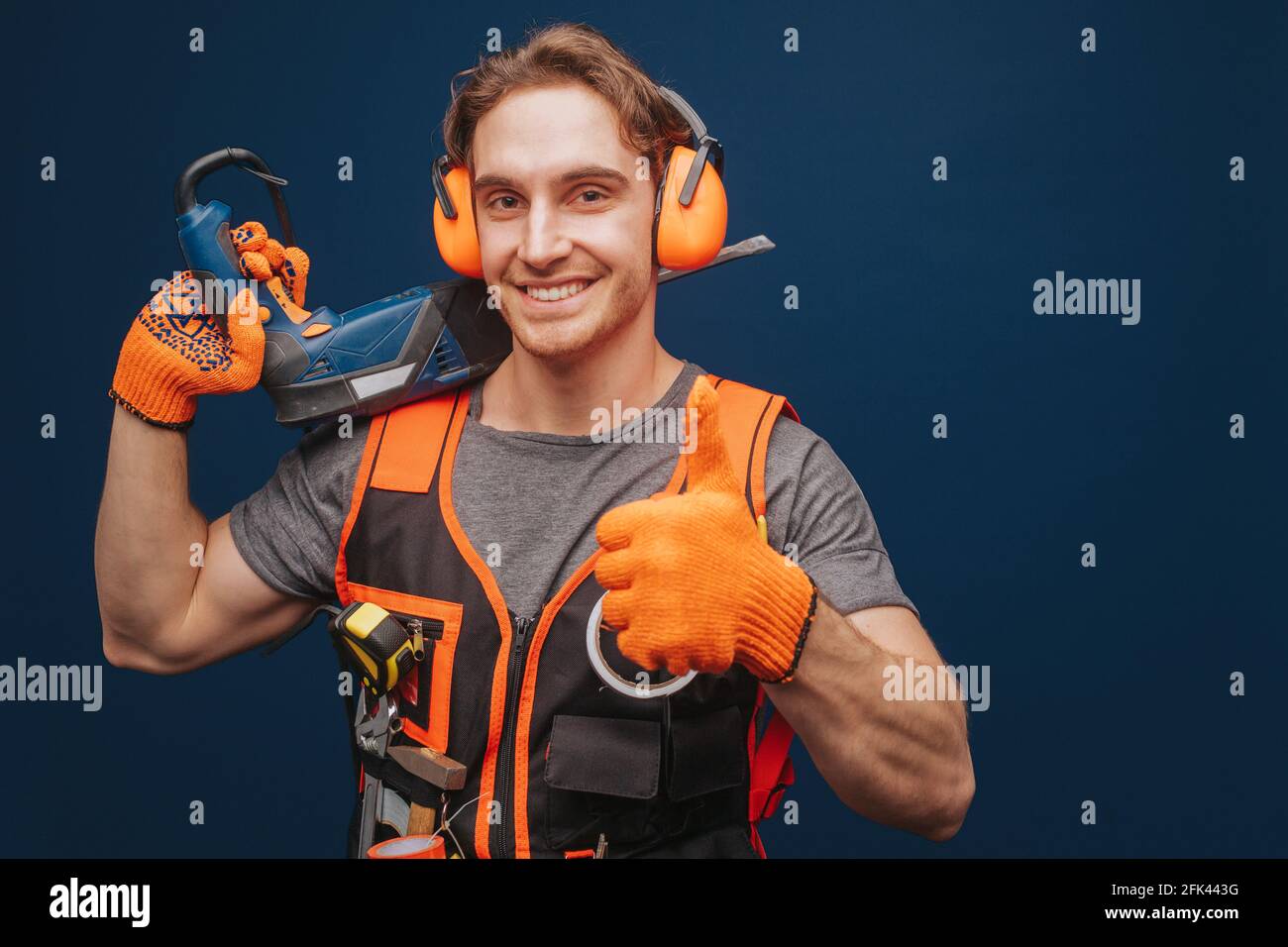 Young builder is showing thumb up while having equipment in arm, isolated over blue background Stock Photo